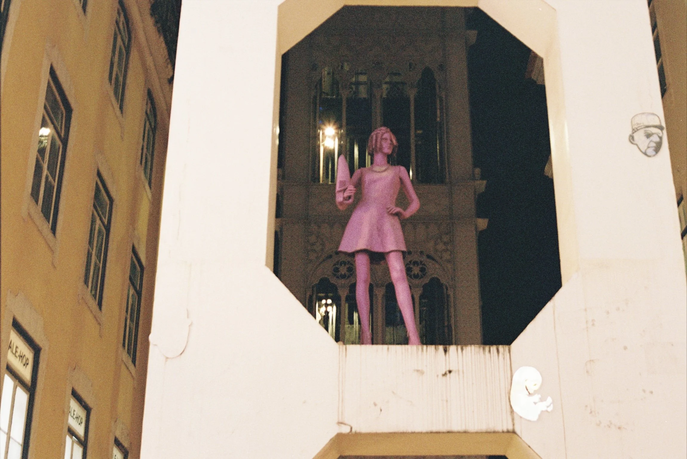 A pink mannequin wearing a vintage-style dress and pearl necklace, standing on a balcony or ledge at night, with the background showing lit windows and the dark exterior of a building.