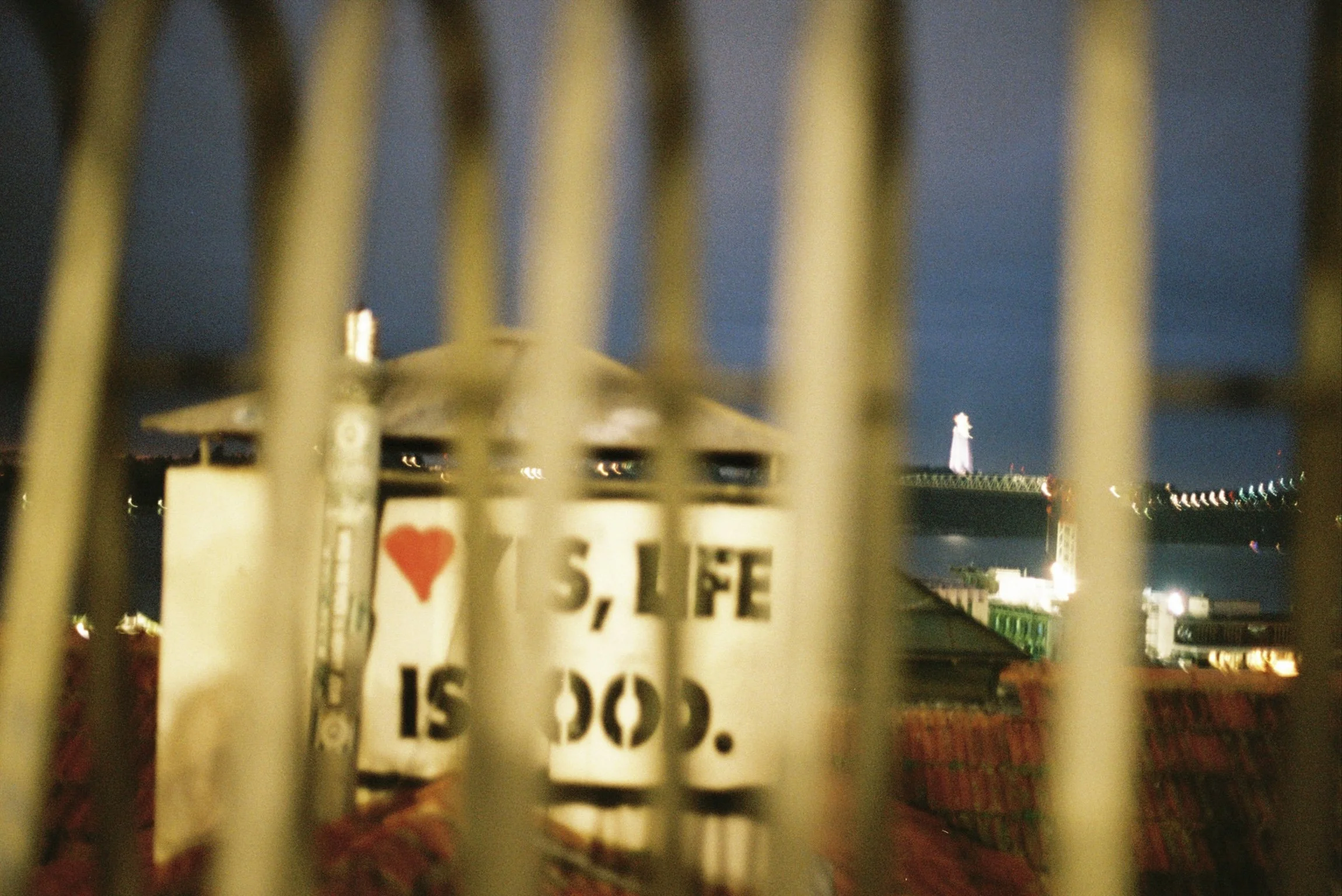 Close-up of a sign with the text 'I Love Seattle' behind a metal fence, with a city skyline and Space Needle visible in the background during nighttime.