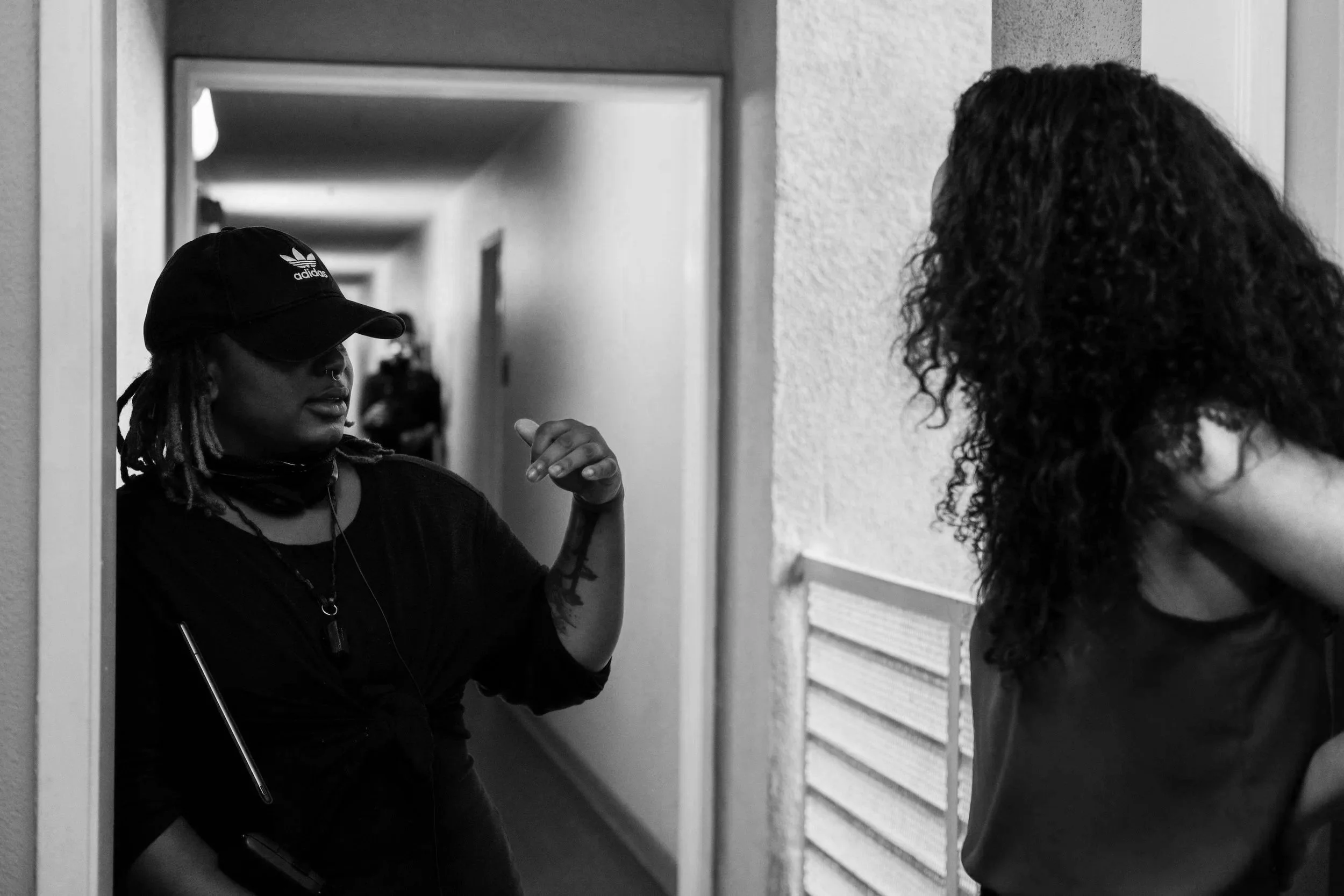 A woman with dreadlocks and a nose piercing, wearing a black Adidas cap, looks into a mirror while a second person with curly hair stands in front of her, in a hallway with textured walls.