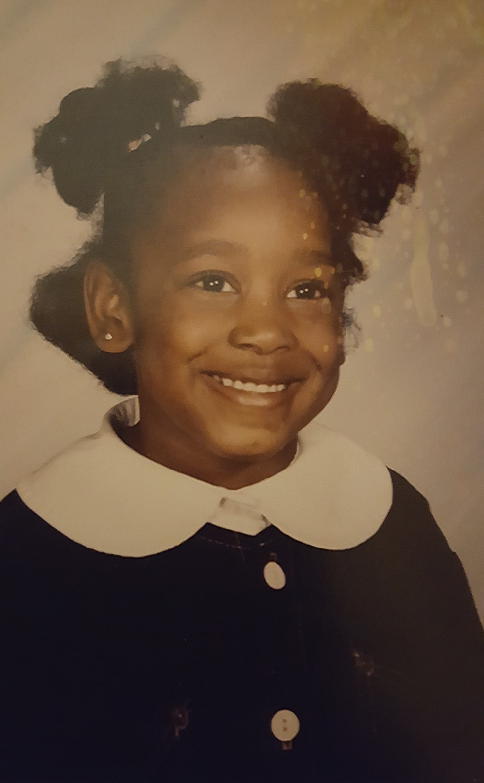 Young girl with dark curly hair styled in two buns, smiling, wearing a black dress with white Peter Pan collar and pearl earrings.