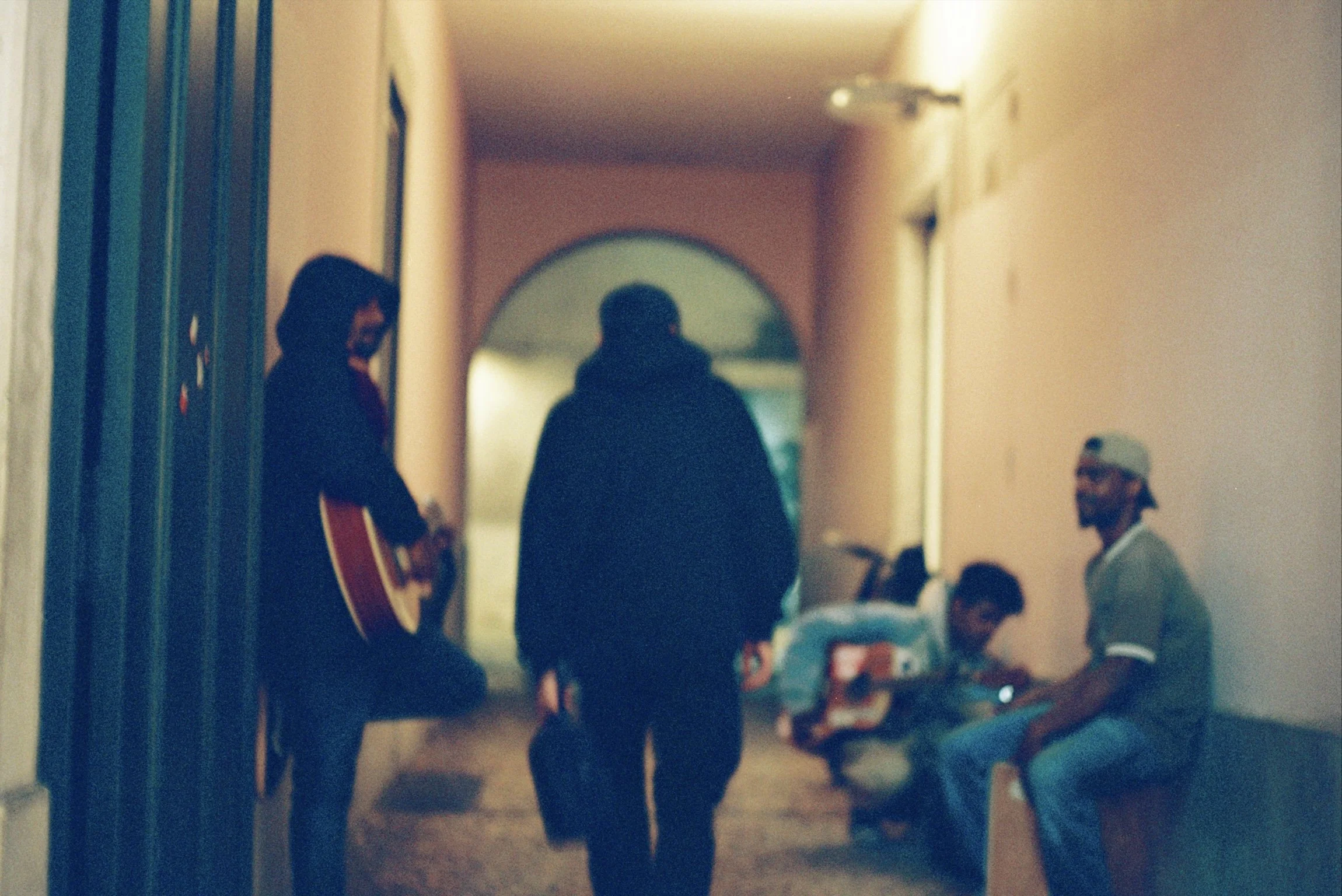 A hallway with people standing and sitting, some with a guitar, and a person playing guitar. The hallway has a doorway at the end with an arch.