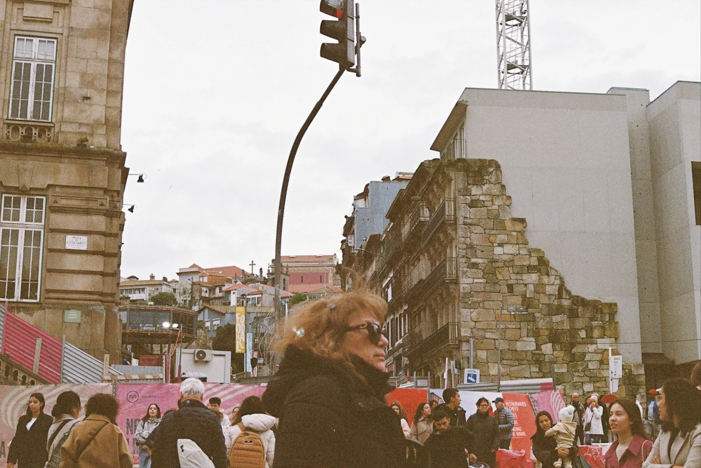 A busy urban scene with many people walking and standing in front of a row of buildings with various architectural styles, including a stone building with ornate window framing and a modern white building. A traffic light is also visible, hanging fro