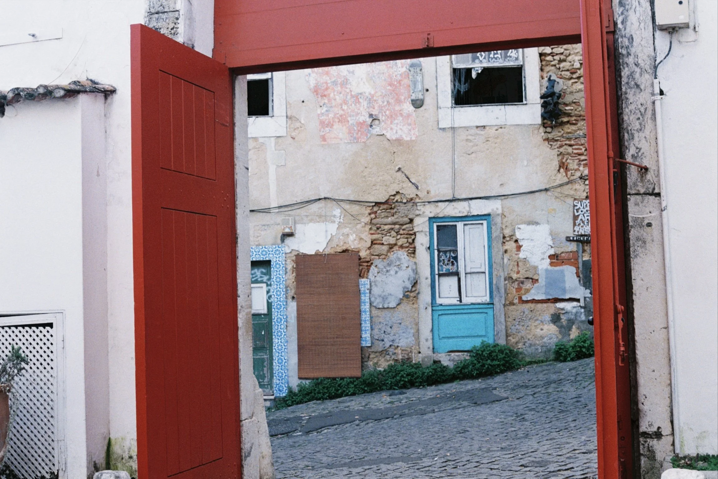 View through a red gate showing a weathered stone and brick building with windows, one framed by blue patterned tiles, and cobblestone street.