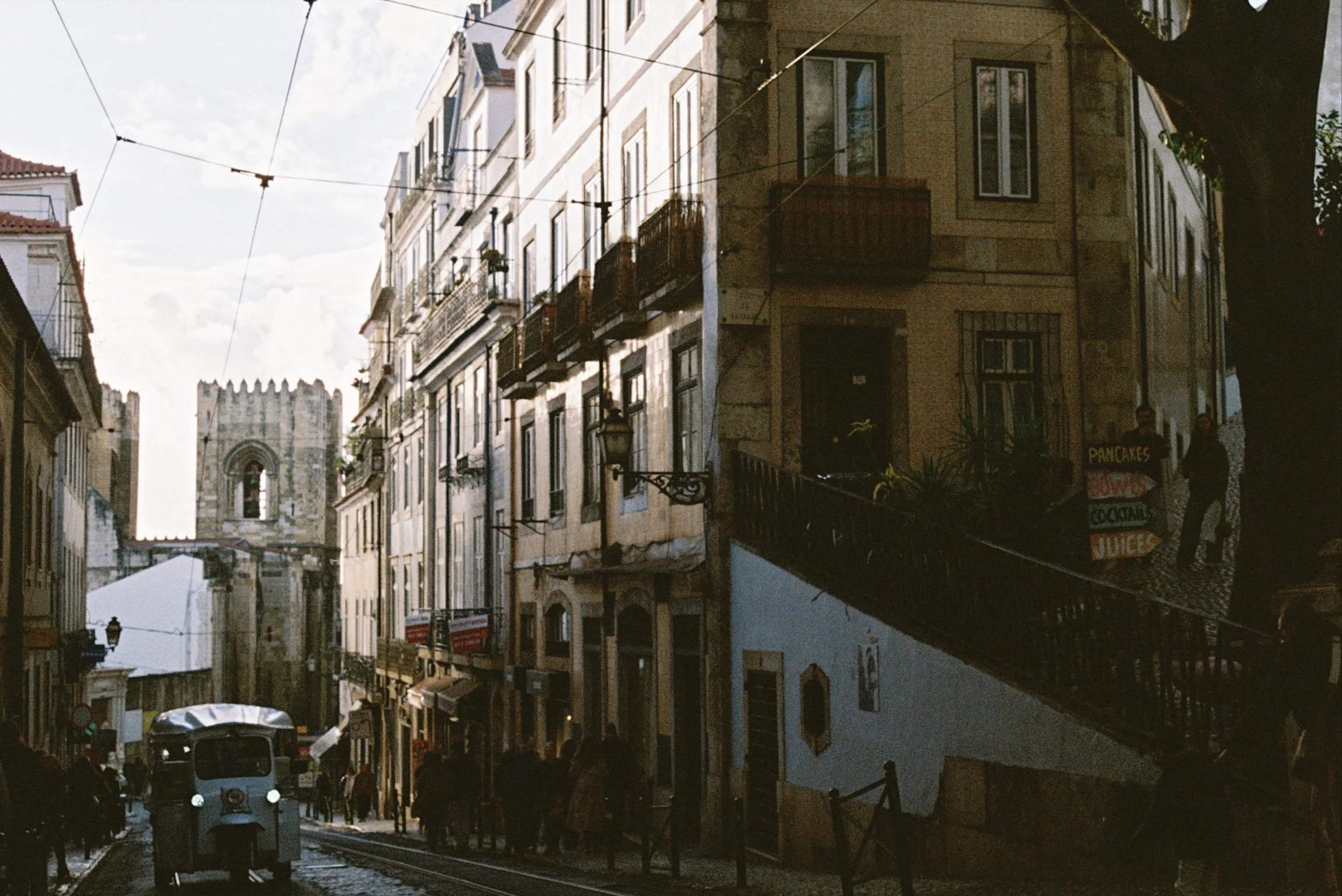 A narrow street with old buildings, some with balconies, and tram tracks in the center. The street is bustling with pedestrians and vehicles, with a historic stone building visible in the background.