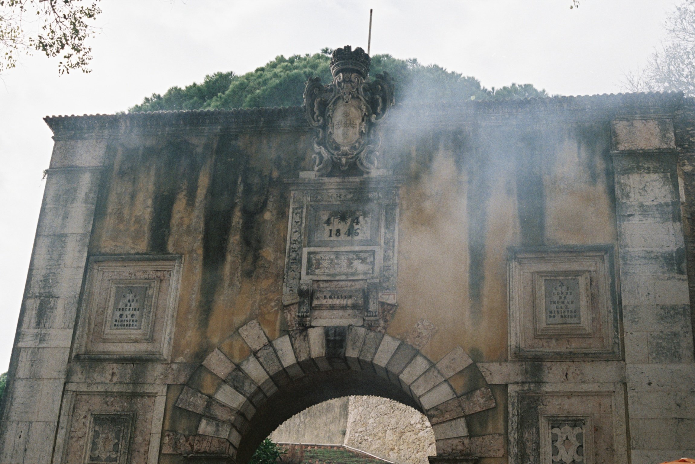 Historical stone archway with a coat of arms and inscriptions, surrounded by trees, showing signs of aging and weathering.