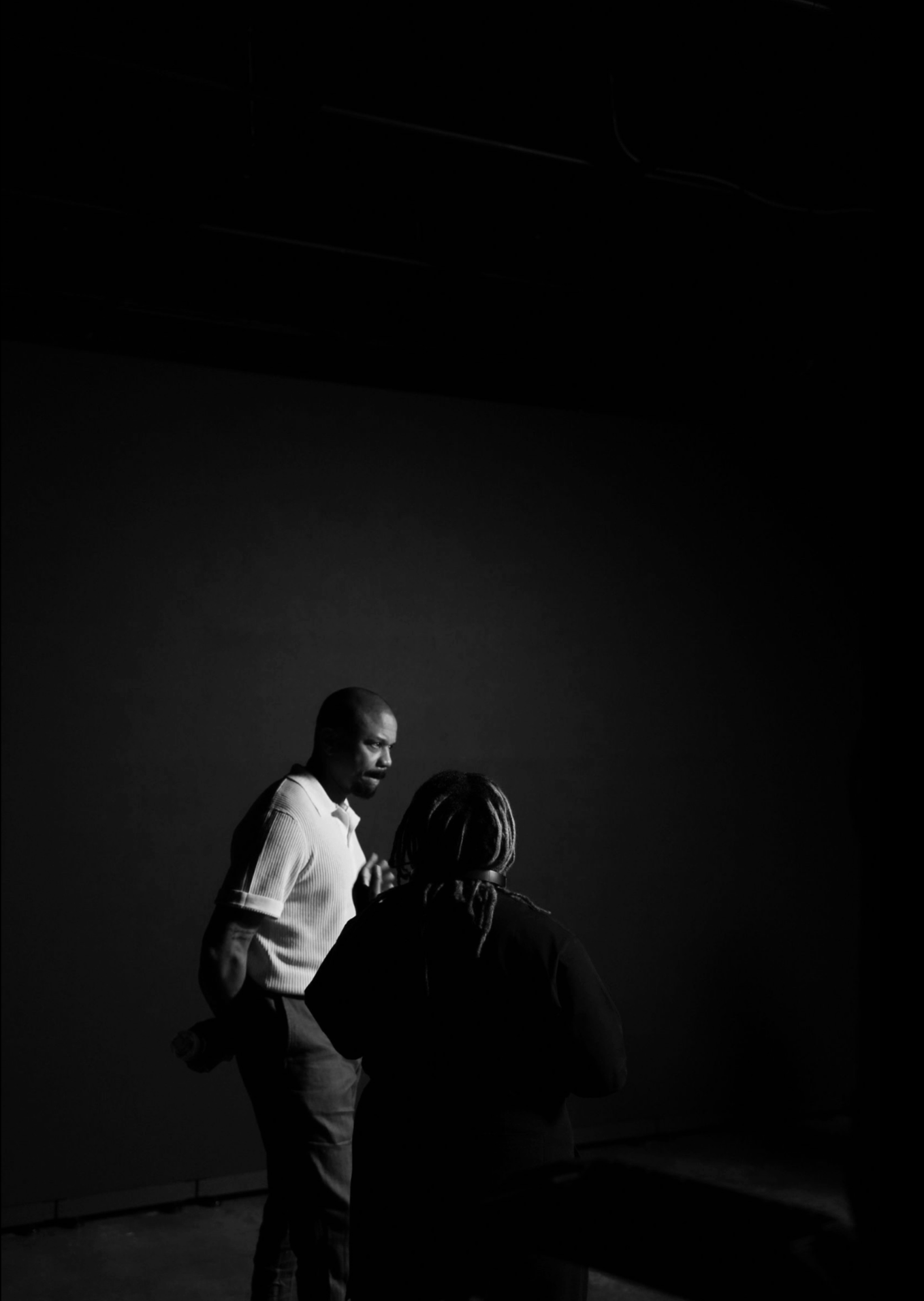 Two men and a woman in a dimly lit room, with one man speaking, the woman listening, and the other man observing in the background.