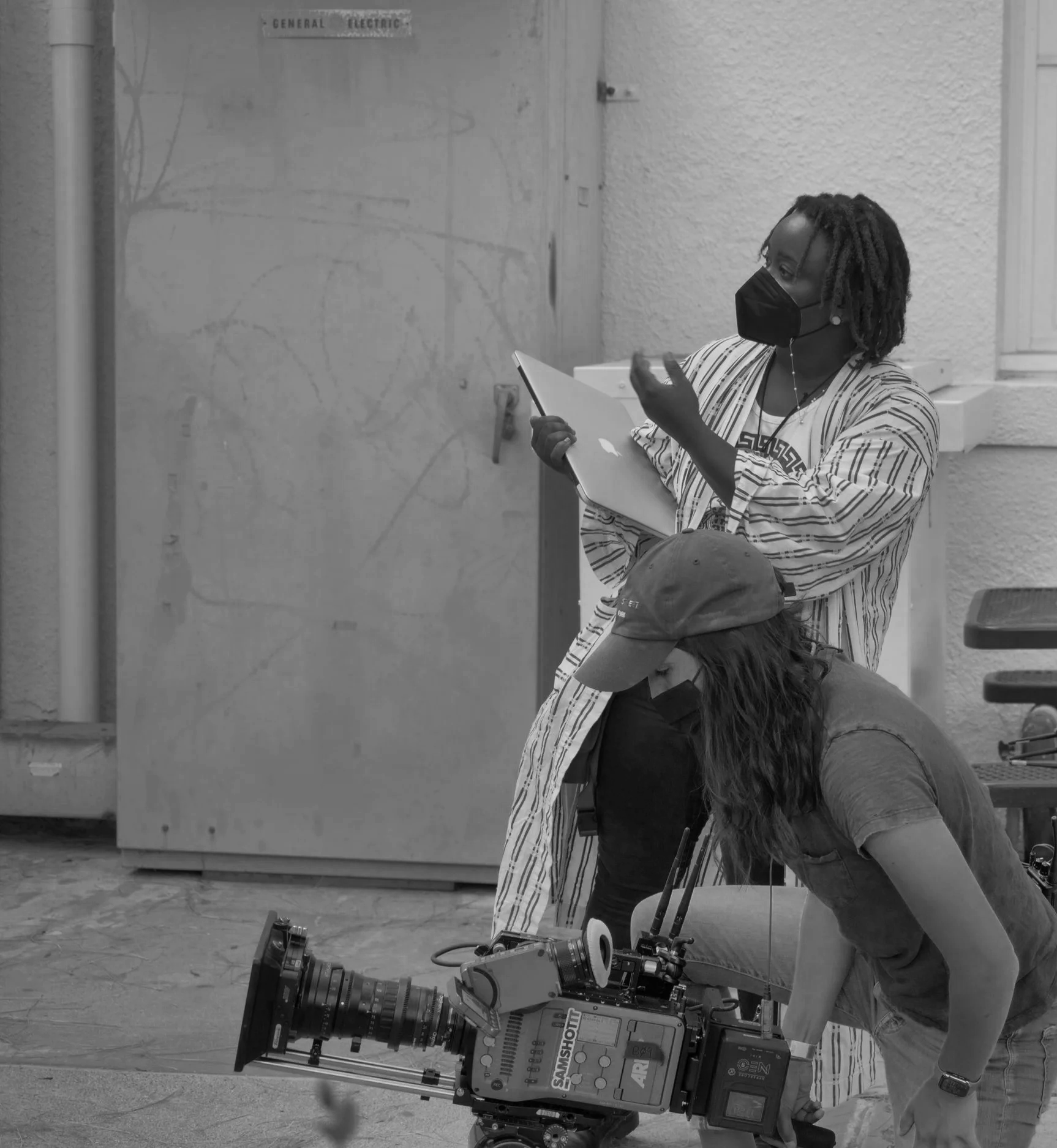 A woman wearing a mask and striped clothing is taking notes on a clipboard. She is standing behind a person operating a professional film or television camera, which is mounted on a tripod. There is a large metal cabinet with a sign that reads 'General Electric' in the background.