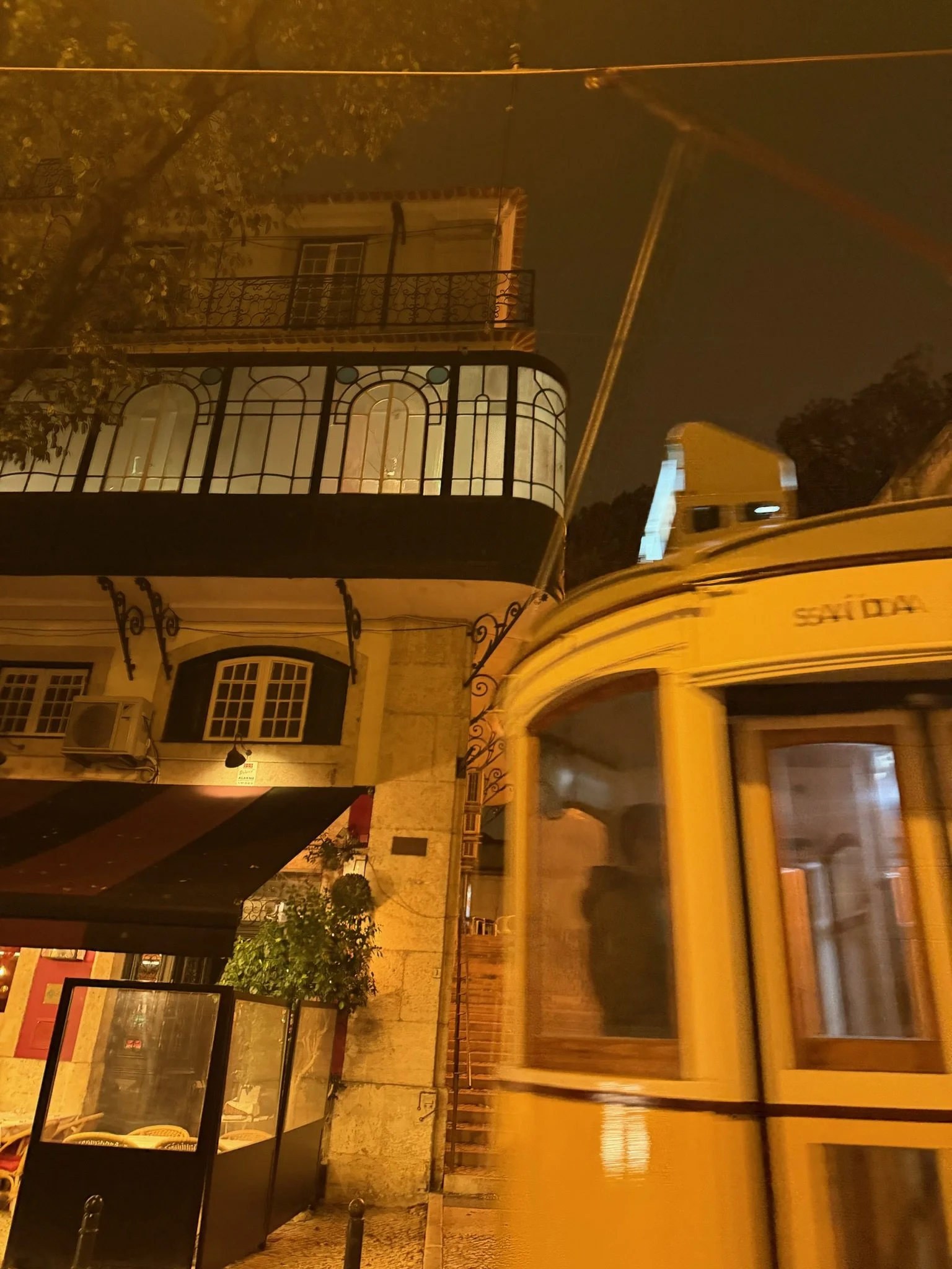 Nighttime city street scene with a yellow city bus passing by a building with balconies and greenery, illuminated warmly.
