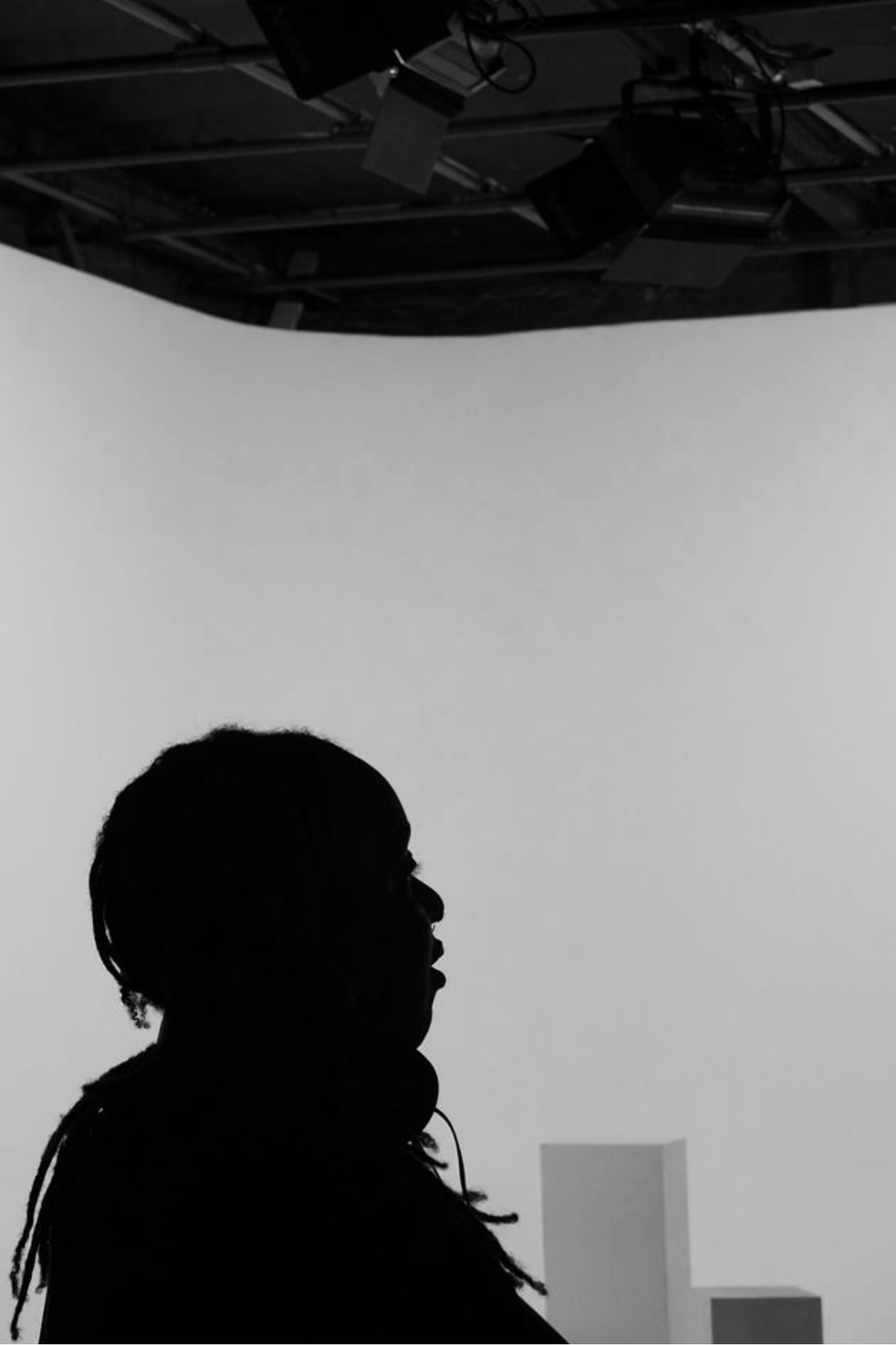 Silhouette of a woman with dreadlocks at a photography or video studio with a rolling black background and studio equipment overhead.
