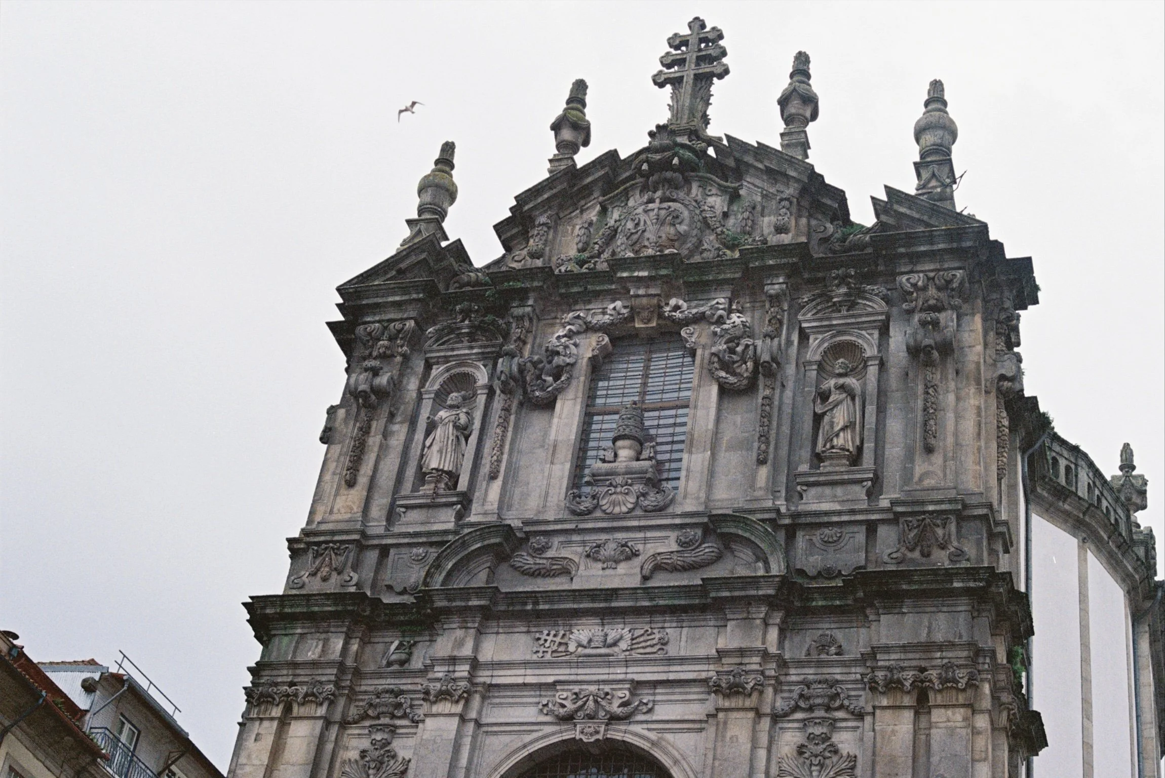 Historical building with ornate stone carvings and statues, topped with crosses, against a cloudy sky, with a bird flying nearby.