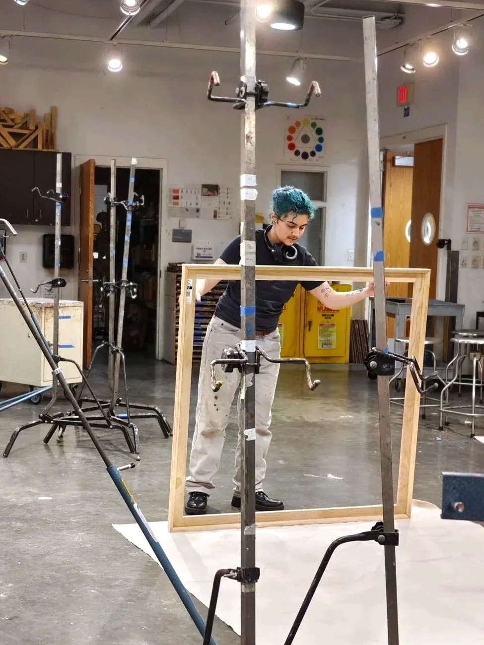 A person with short blue hair working on a large wooden frame in a workshop. The person is holding the frame while standing behind it, surrounded by various tools and equipment, with white walls and bright overhead lights.