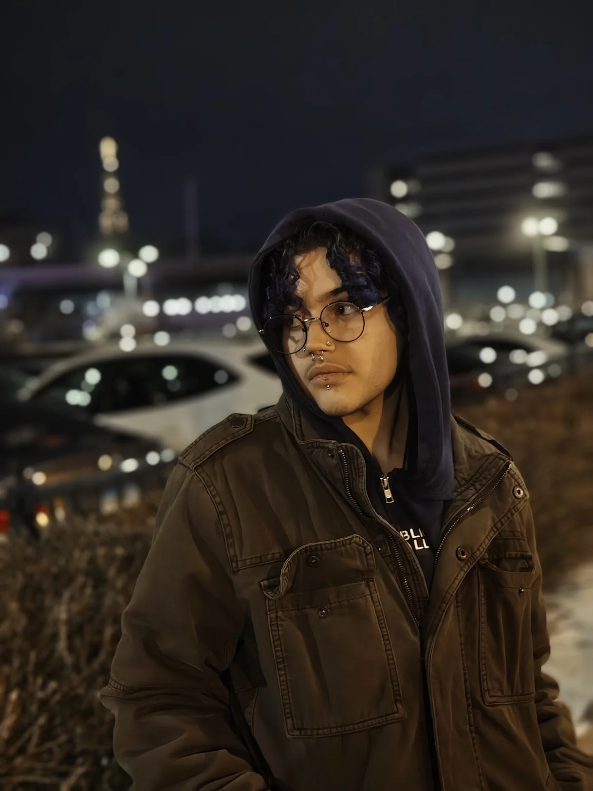 A young man with curly black hair, glasses, and facial piercings, wearing a hoodie and a jacket, stands in a parking lot at night with city lights and cars in the background.