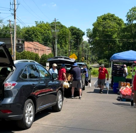 People at an outdoor market with cars parked and a tent distributing food on a sunny day.