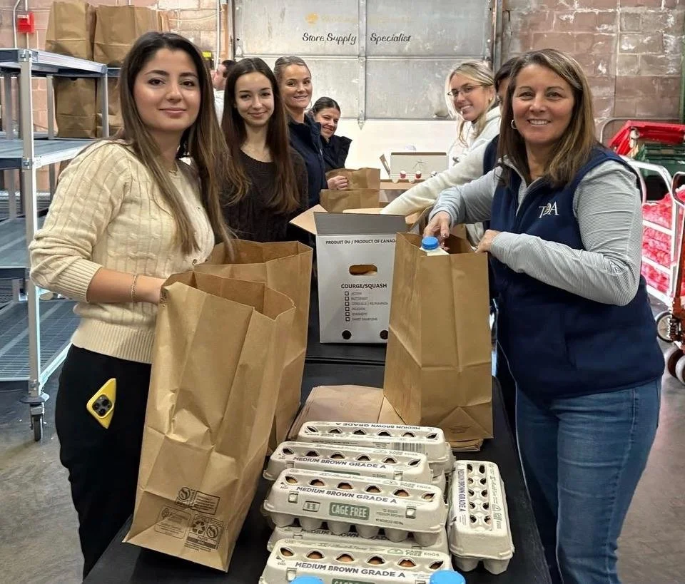Group of six women standing at a table with brown paper bags and cartons of eggs, smiling. They are in a warehouse or distribution center.