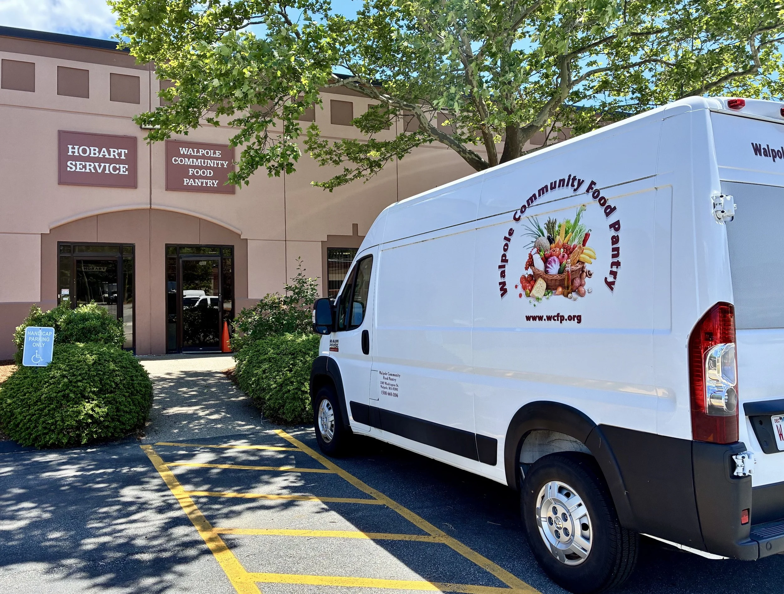 A white Walpole Community Food Pantry delivery van parked. The building has two large signs, one reading 'HOBART SERVICE' and the other 'WALPOLE COMMUNITY FOOD PANTRY'. There is a handicap parking sign, greenery, and a large tree with green foliage.