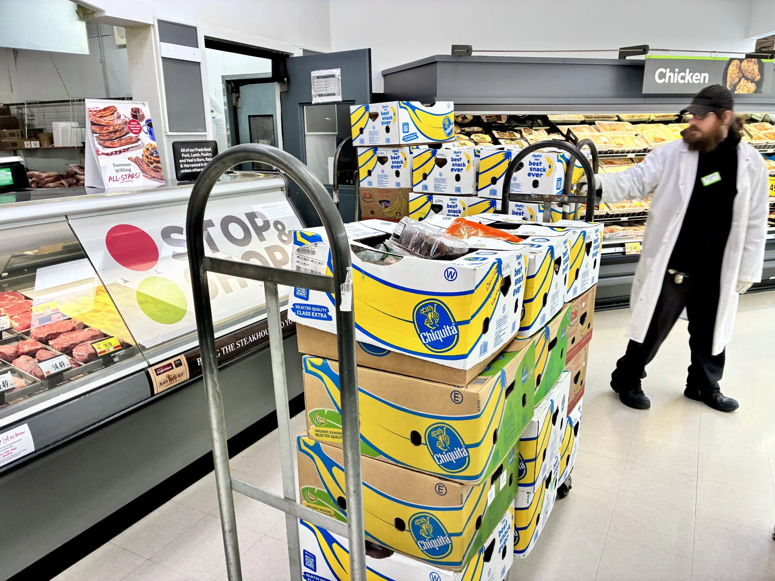A man shopping at a grocery store, reaching for a cart near displays of bananas and other produce, with frozen chicken products visible on the shelves behind him.