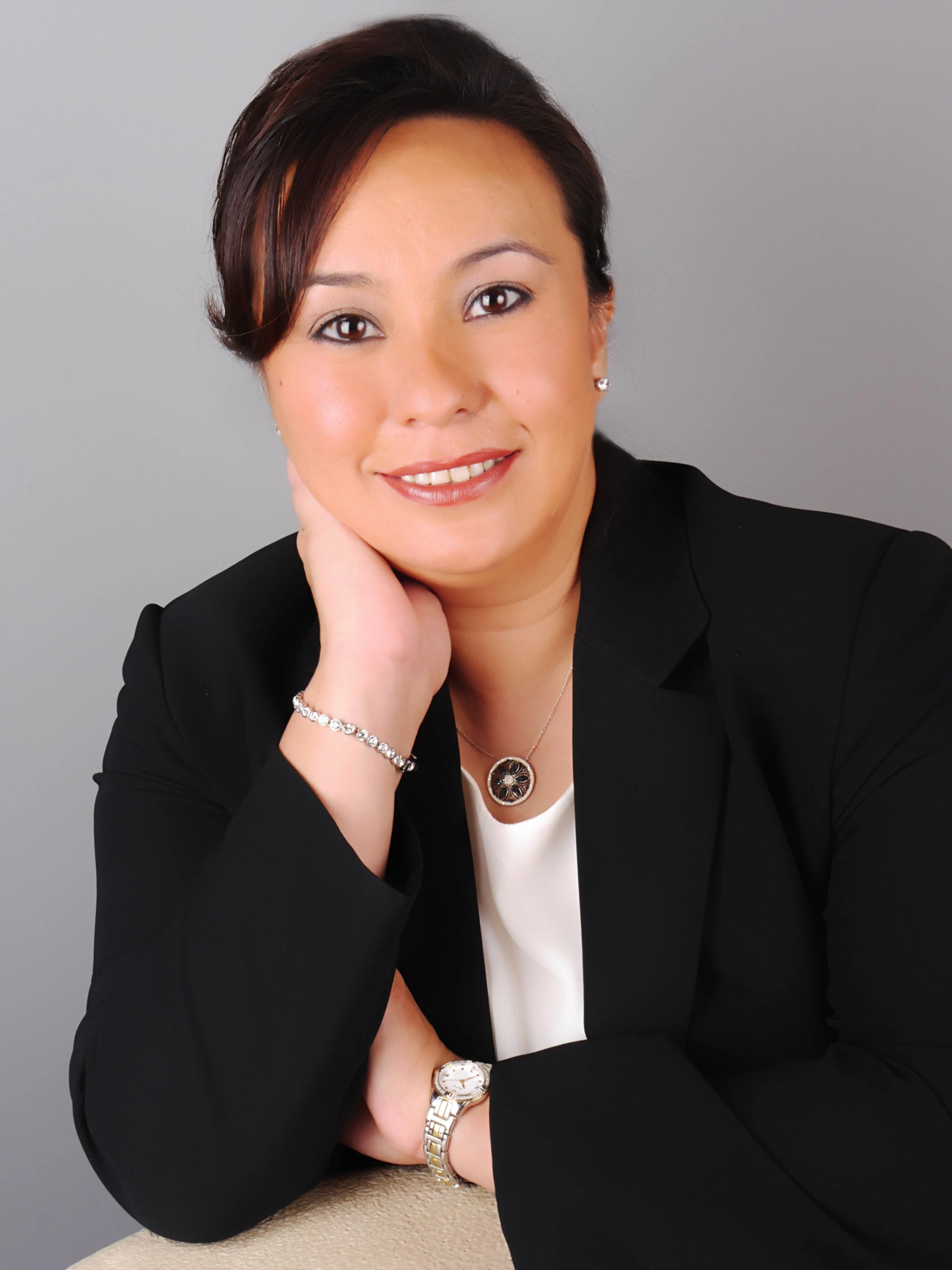 A woman with short dark hair, wearing a black blazer, white top, jewelry including earrings, a bracelet, a watch, and a necklace, posed with her head resting on her hand, smiling softly against a neutral gray background.
