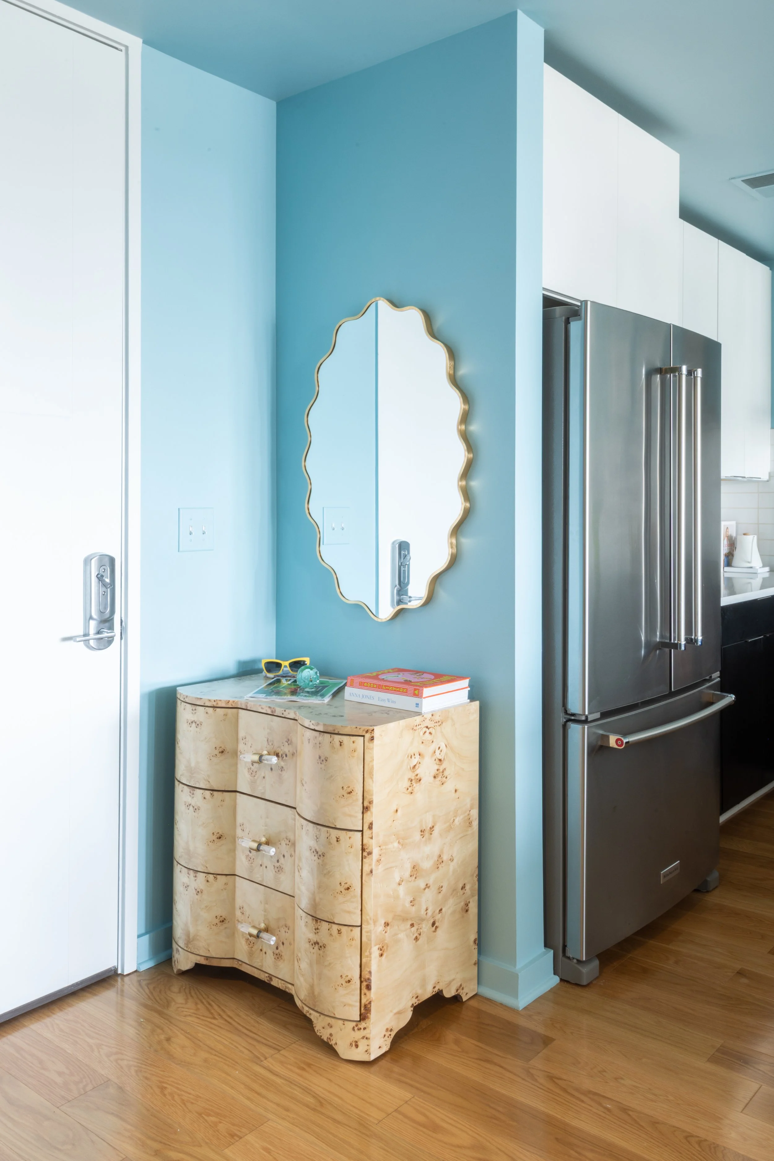 A cream-colored wooden dresser with a curved front and small drawers with gold handles, standing next to a blue wall in a modern kitchen. On top of the dresser are a pair of yellow sunglasses, a small turquoise decorative object, and books. Behind the dresser, a mirror with a gold wavy frame is mounted on the wall. To the right, a stainless steel refrigerator and part of a white countertop are visible, with wooden flooring throughout the space.