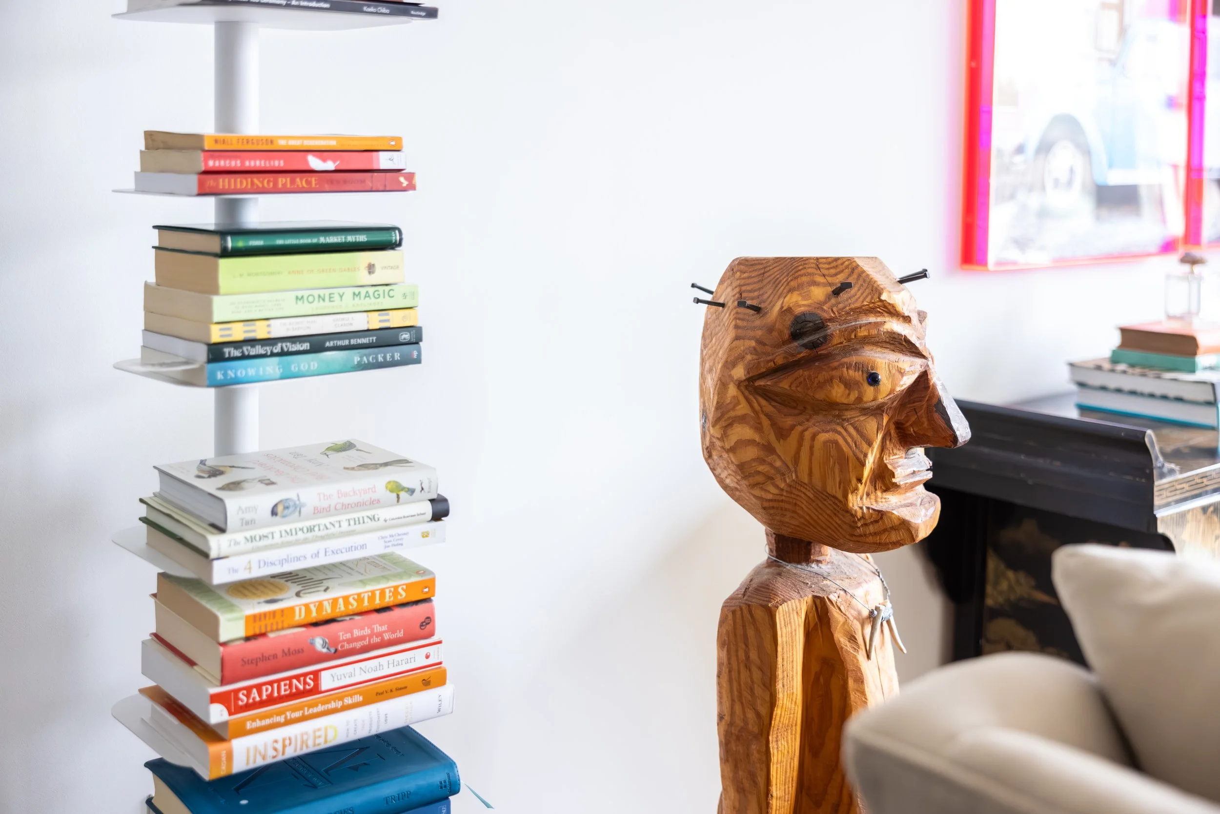 Books stacked on a white rotating shelf with various titles, a carved wooden sculpture of a person's head, part of a colorful framed artwork in the background, and a black table or desk nearby.