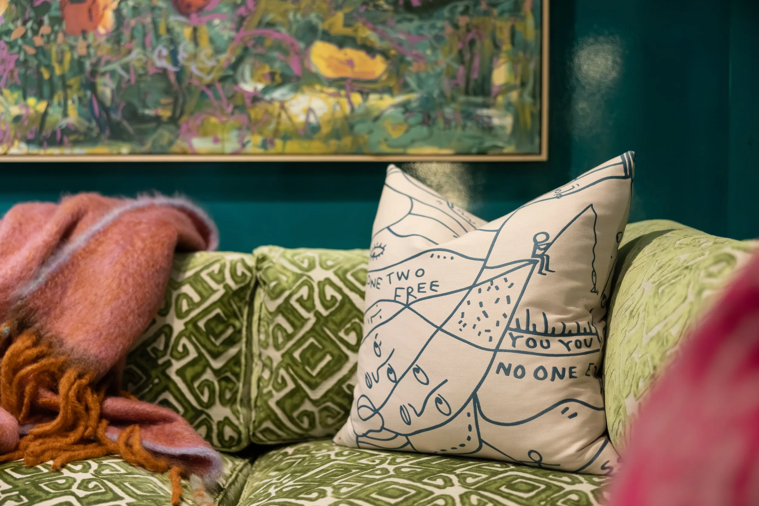 Close-up of a cozy seating area with a green patterned cushion, a white pillow with a map and text, a green pillow, a pink fleece blanket, and a multicolored abstract painting on the wall in the background.