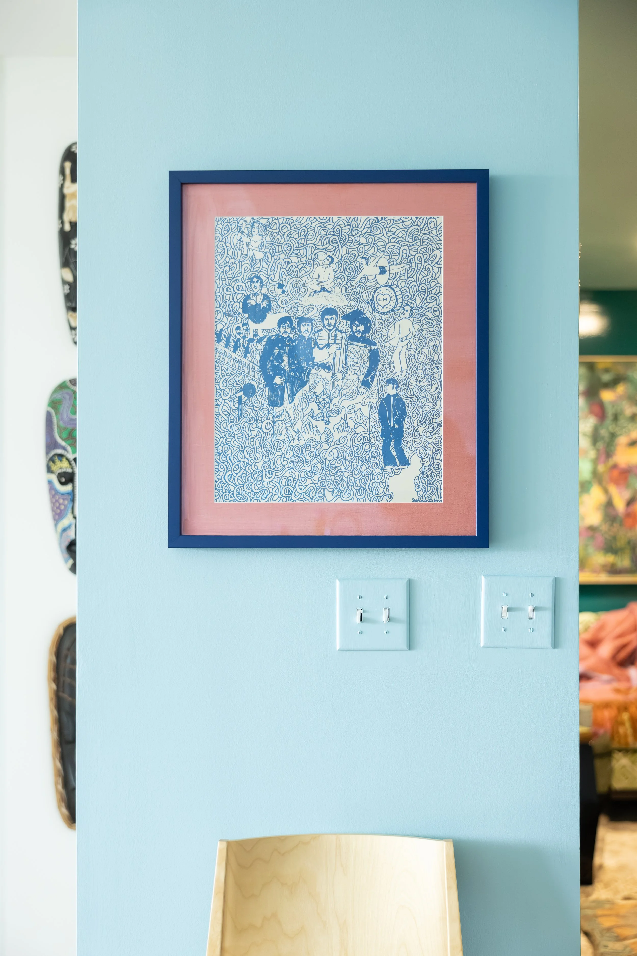 A framed drawing hangs on a light blue wall, with a pink mat behind it. The drawing features multiple people, abstract patterns, and intricate line work, created in blue ink.