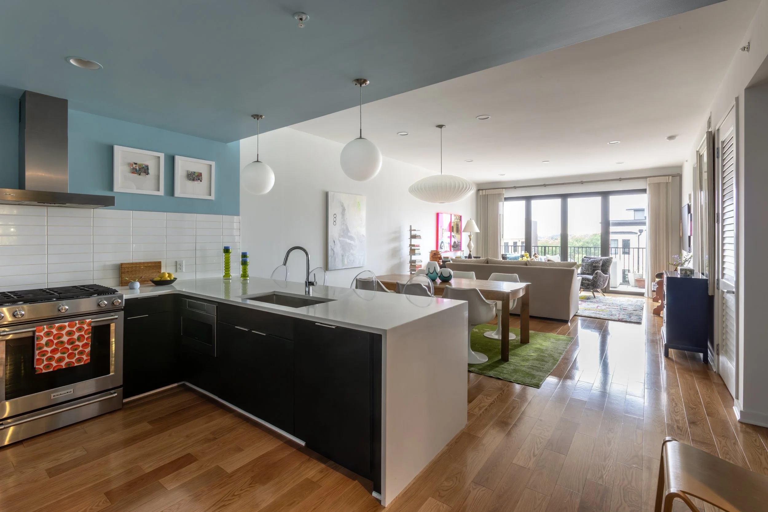 Open-concept living room and kitchen with hardwood floors, white and blue walls, large windows, and modern decor.