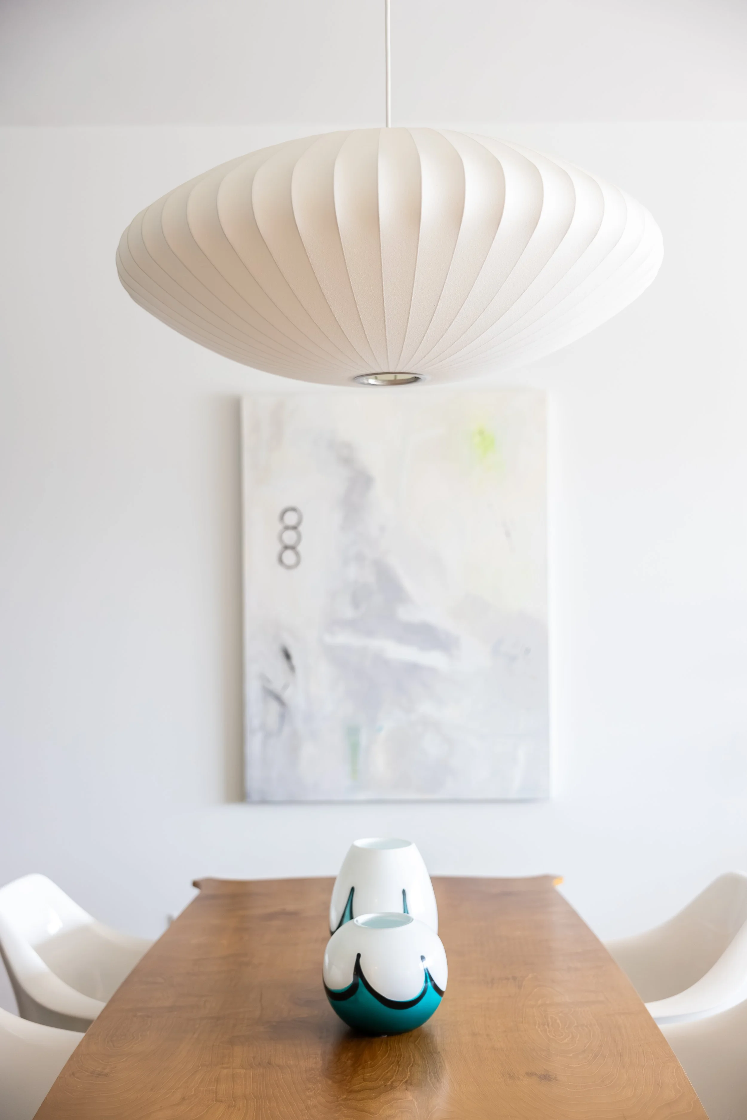 A modern dining room with a wooden table, white chairs, three decorative vases on the table, a large abstract painting on the wall, and a white paper pendant light hanging from the ceiling.