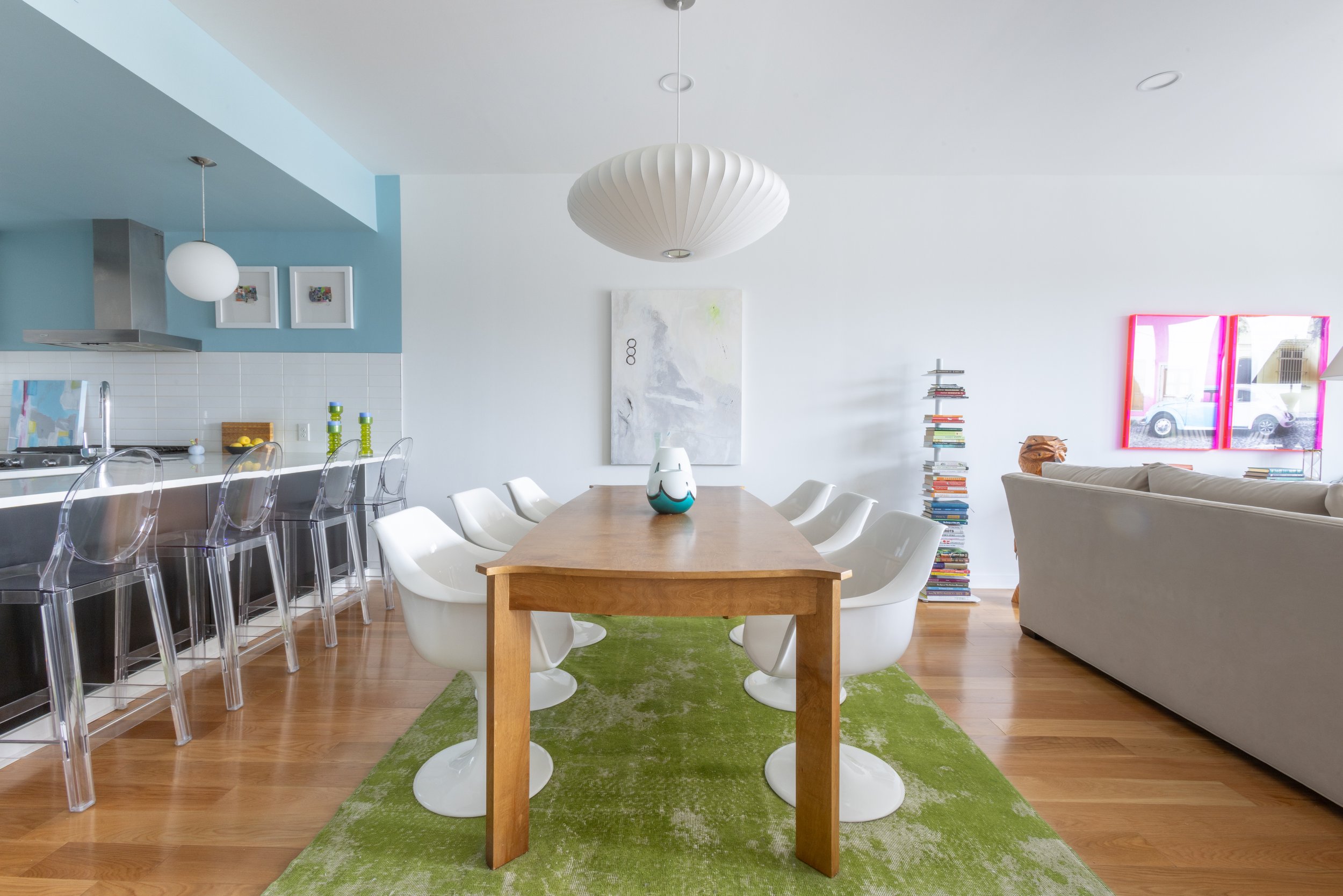 A dining area with a wooden table and white chairs on a green rug, a kitchen with a blue accent wall, hanging lights, a wall with art, a bookshelf with stacked books, and a bright window.