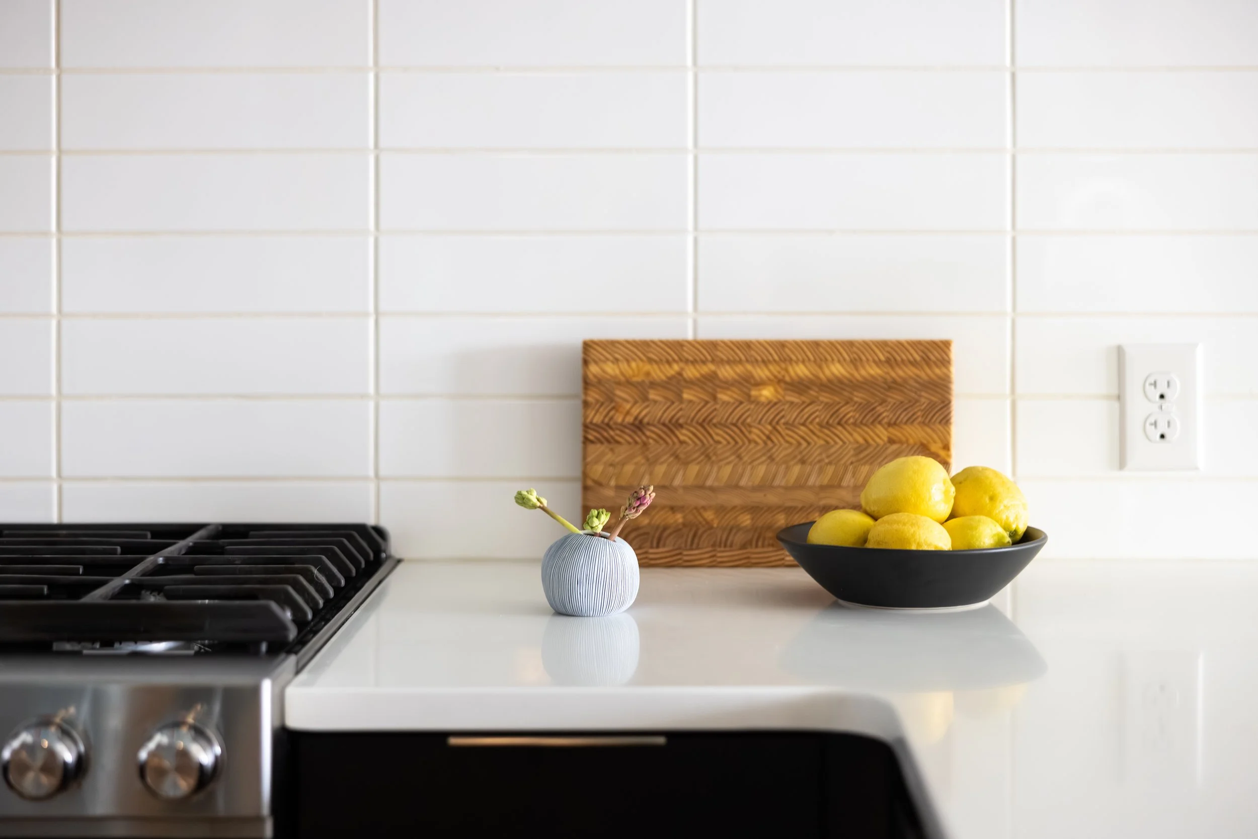 A kitchen countertop with a black bowl filled with lemons, a small vase with a flower, a wooden cutting board, and a white backsplash with an electrical outlet.