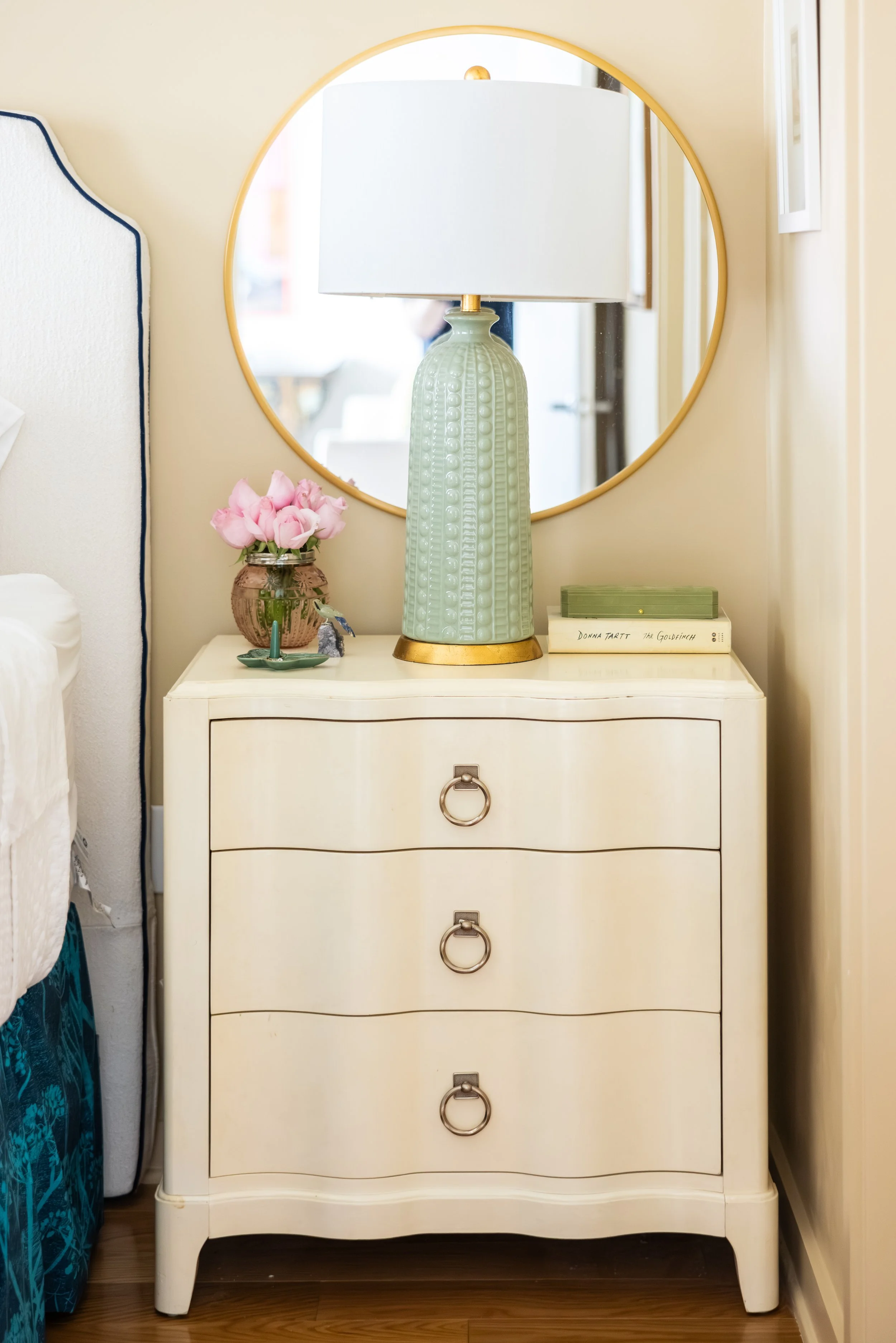 Cream-colored dresser with three drawers and large ring pulls, sitting on a wooden floor, with a tall ceramic lamp with a white shade, pink flowers in a copper vase, and hardcover books on top, and a round mirror above it reflecting a window and part of a room.