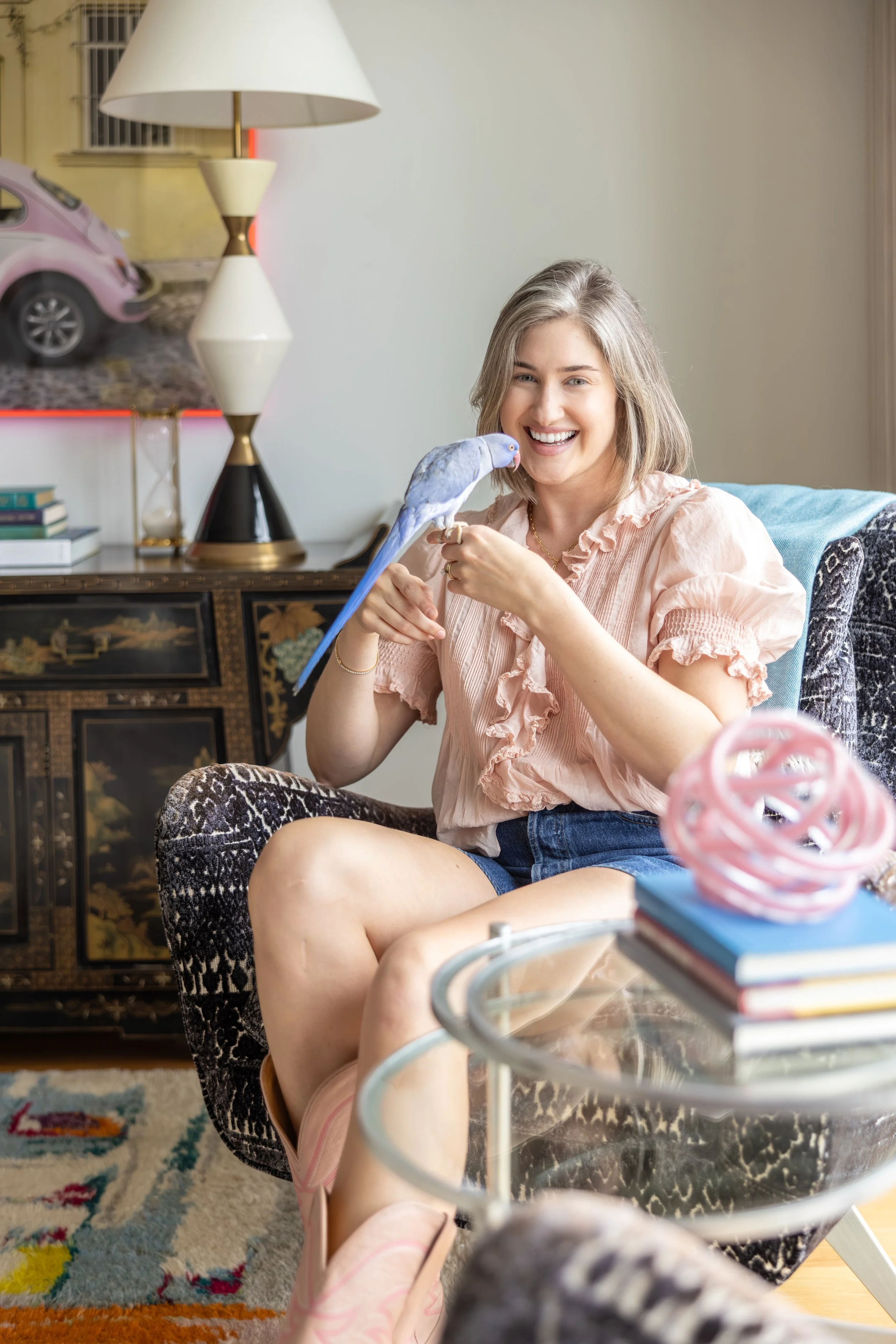A smiling woman with gray hair sitting on a patterned chair, holding a blue and purple bird on her finger, in a cozy living room with a colorful rug, table, and books.