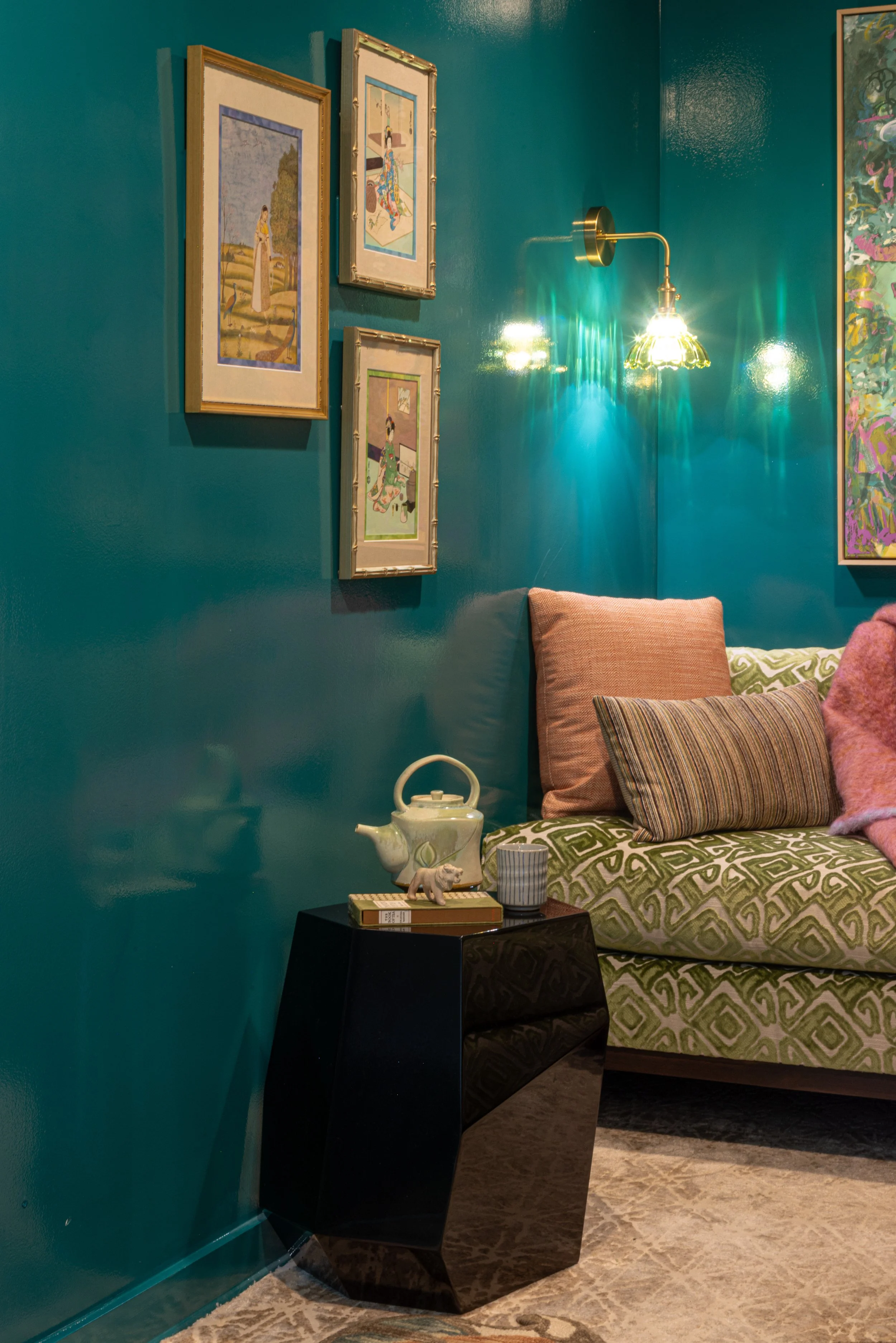 A corner of a room with teal walls decorated with framed artwork. A wall-mounted lamp illuminates the area. There is a bed with patterned green and beige bedding and a variety of pillows. A black bedside table holds a white teapot with a small animal figurine, a striped mug, and a book. The floor is carpeted with a patterned rug.