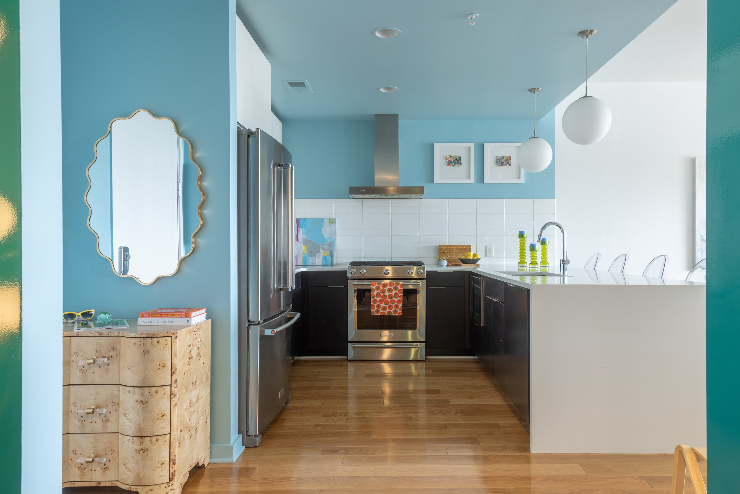 A modern kitchen with blue walls, white tile backsplash, stainless steel appliances, black cabinets, and a white countertop. Decor includes three round pendant lights, two framed artworks, a bowl of lemons, green glass bottles, and a mirror with a gold frame. There's a natural wood floor and a small beige side table with books and sunglasses.