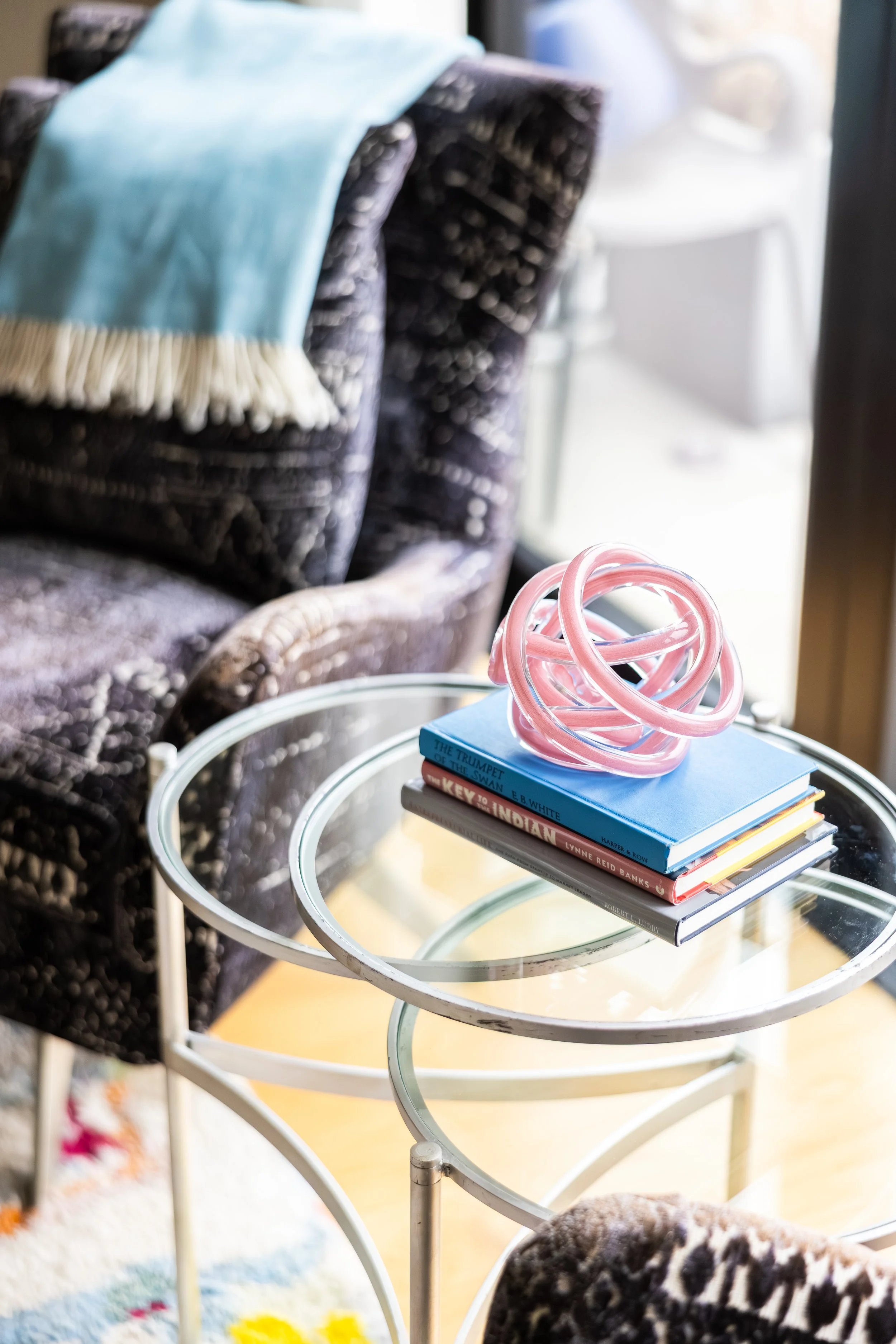 A stack of books on a glass-top side table, with a pink decorative object on top, in a cozy room with patterned furniture and sunlight coming through a window.