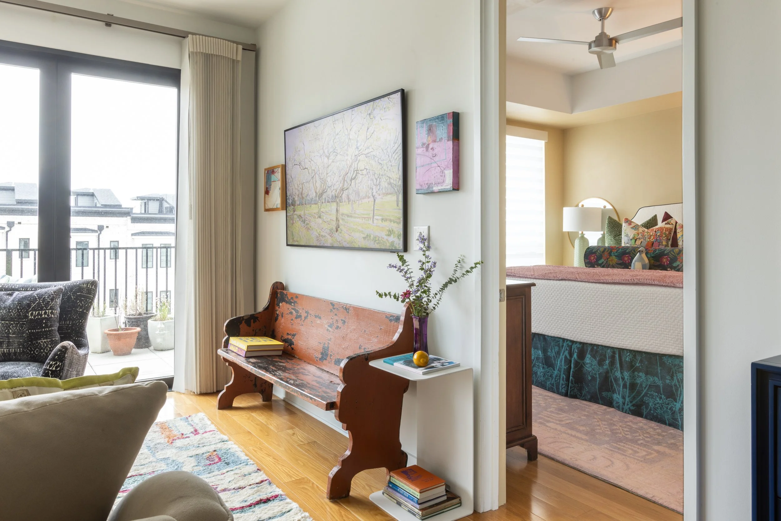 Interior view of a contemporary apartment with a living room and bedroom. The living room has large windows with beige curtains, a colorful patterned rug, and modern furniture including a white armchair and black patterned chairs. There is a wooden bench, a white side table with books and a yellow decorative object, and a vibrant floral arrangement in a purple vase. The wall features framed artwork and a small plant. The bedroom, visible through an open doorway, includes a bed with a white headboard, colorful pillows, a vanity mirror, and a bedside lamp. A ceiling fan is installed in the bedroom.