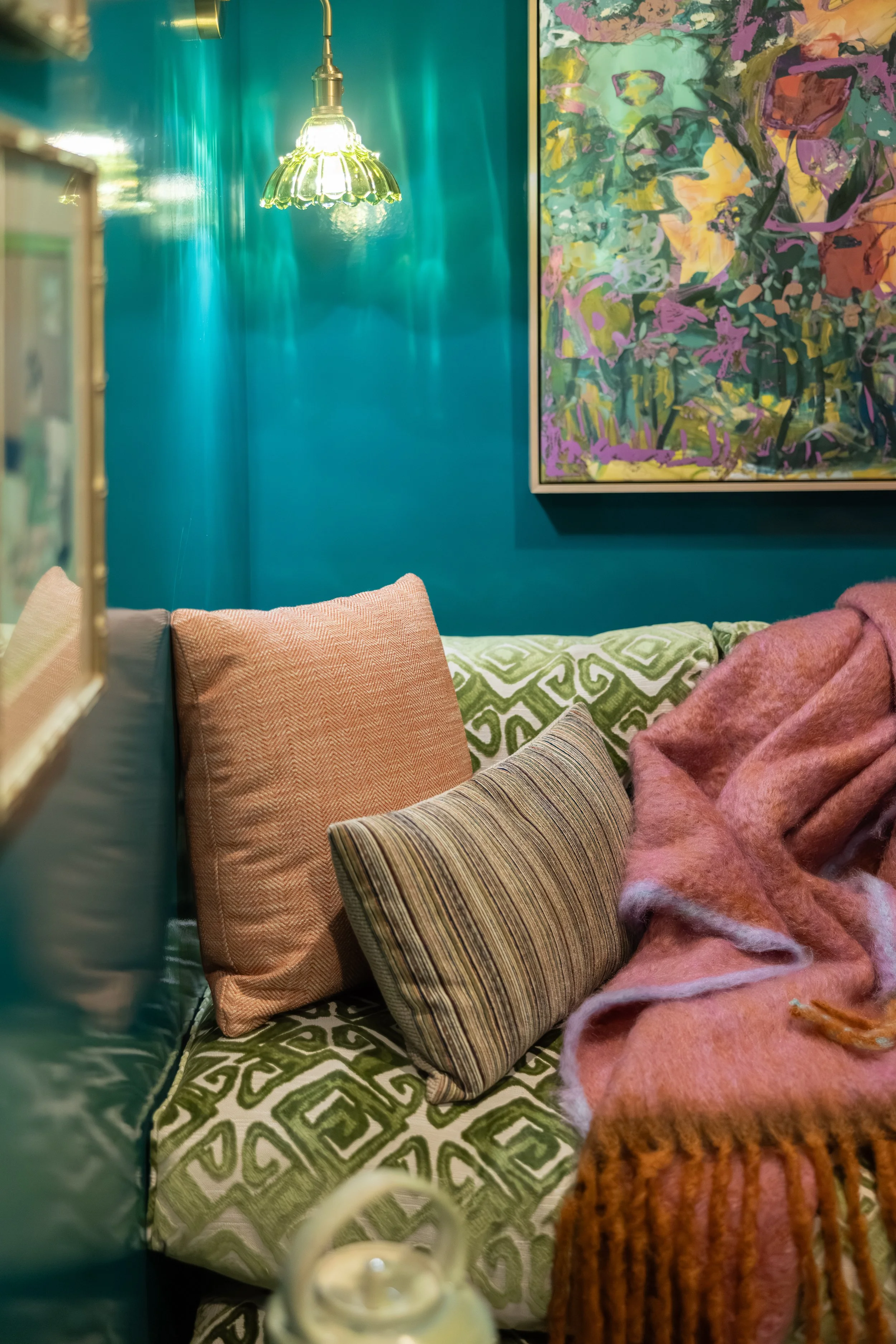 A cozy corner with colorful pillows on a patterned green and white blanket. A pink fuzzy blanket is draped over the cushions, and a painting with vibrant abstract colors hangs on the turquoise wall. There is a brass pendant light hanging from the ceiling.