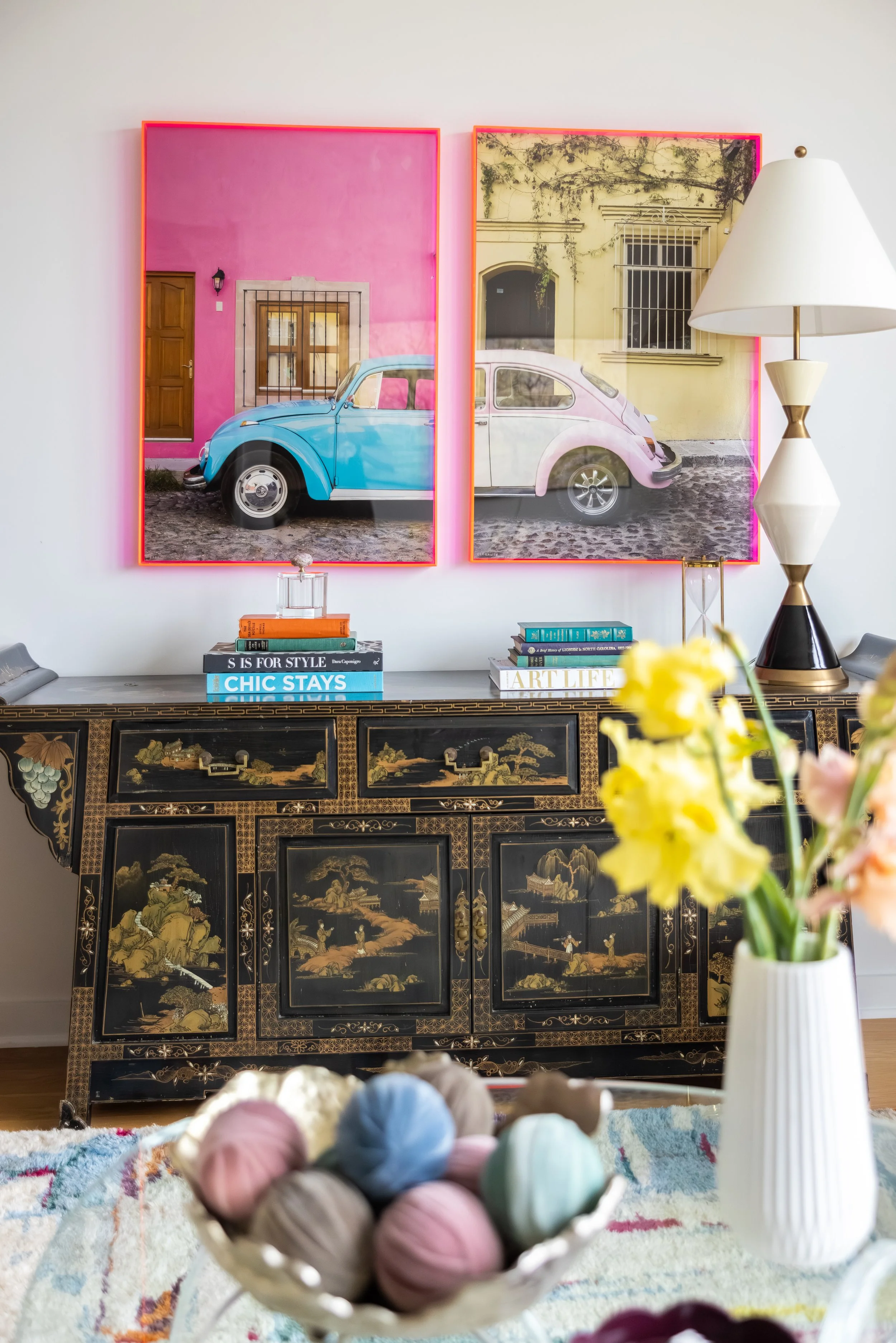 Colorful artwork featuring two vintage Volkswagen Beetles, one painted in bright blue and the other in soft pink, displayed against a backdrop of pastel-colored buildings in a cityscape.