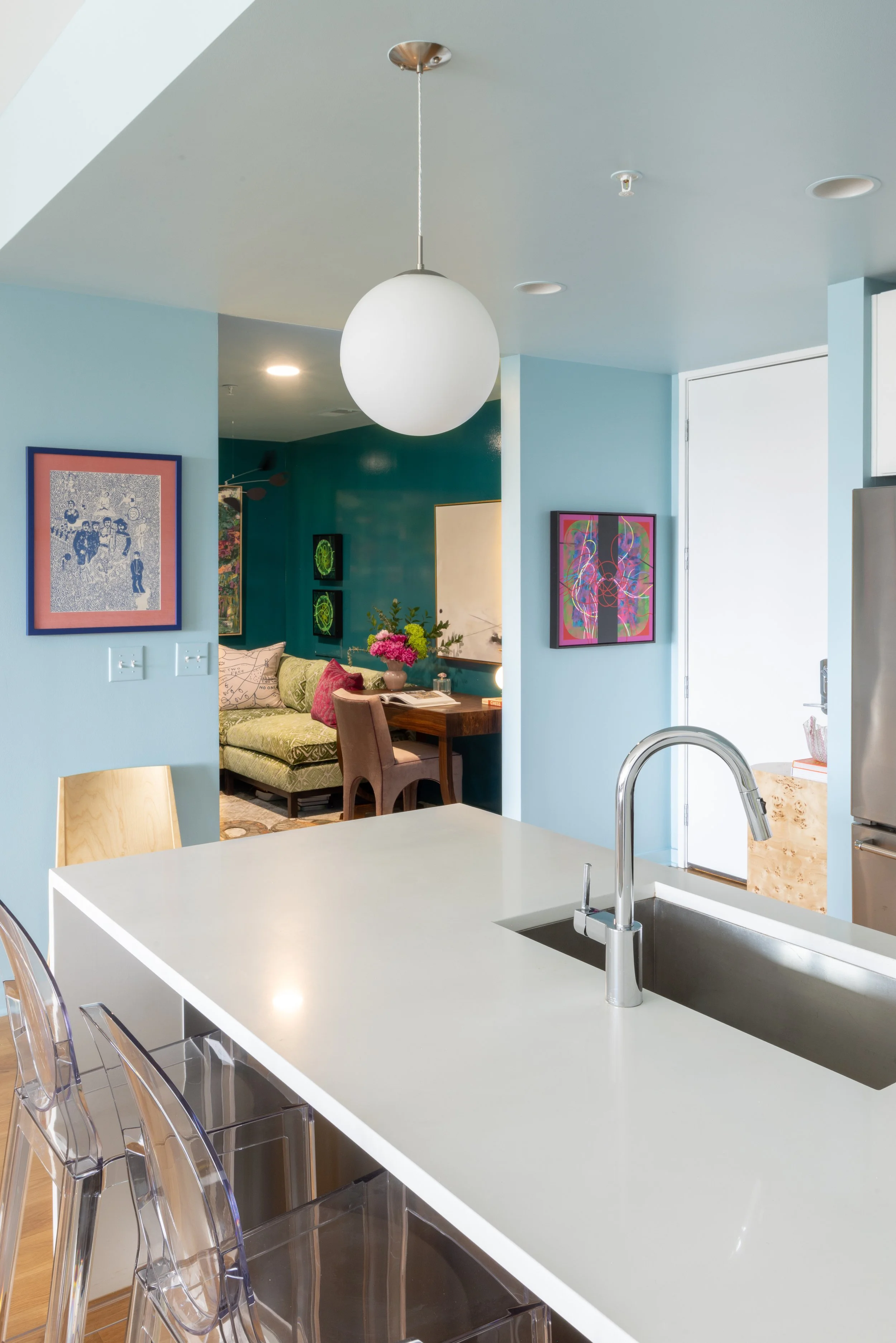 Contemporary kitchen with white island, transparent chairs, light blue walls, and artwork in the background, visible through an open space.