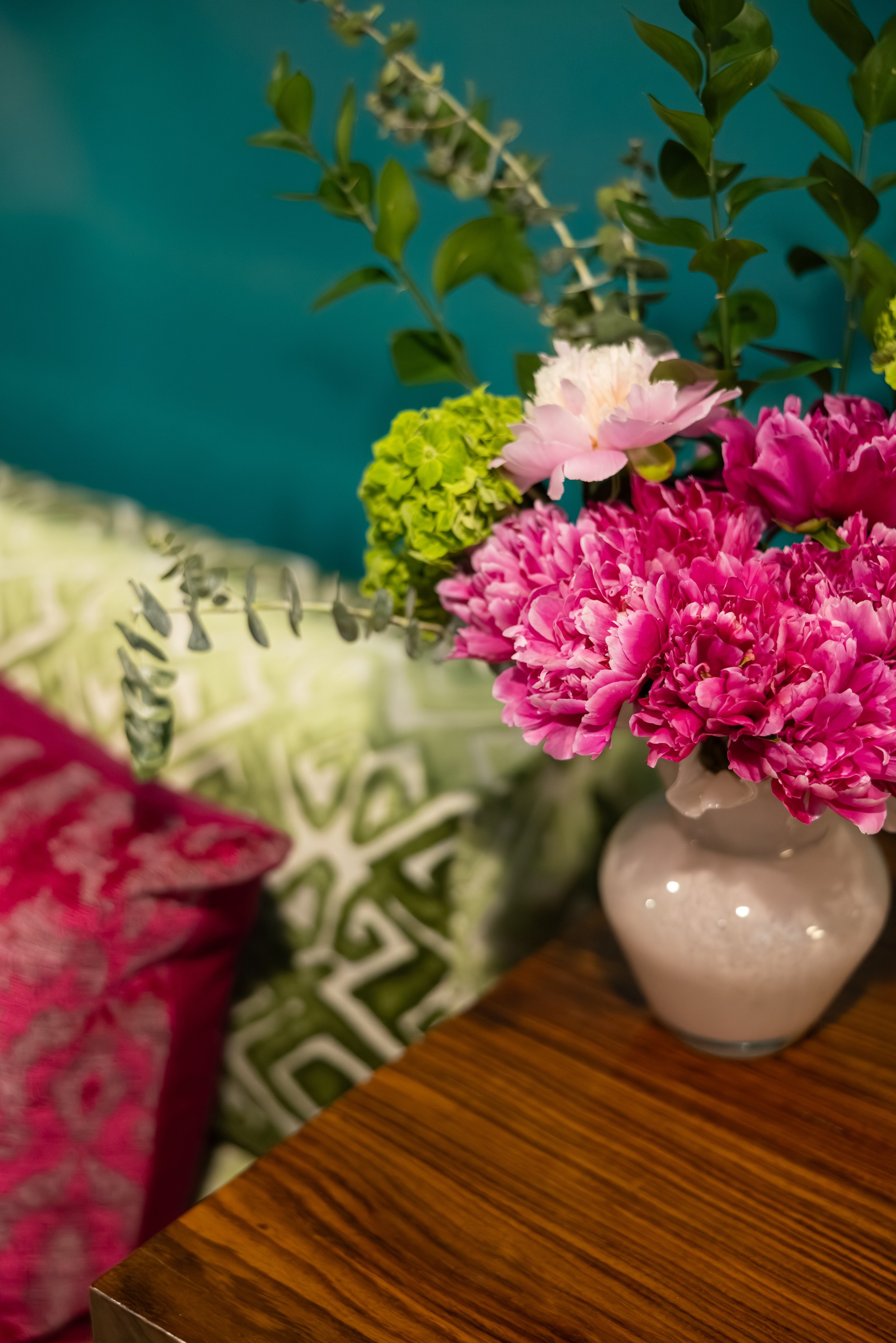 A pink and white floral arrangement in a white ceramic vase on a wooden table, with part of a green and white patterned cloth, and a pink floral patterned cloth in the background.