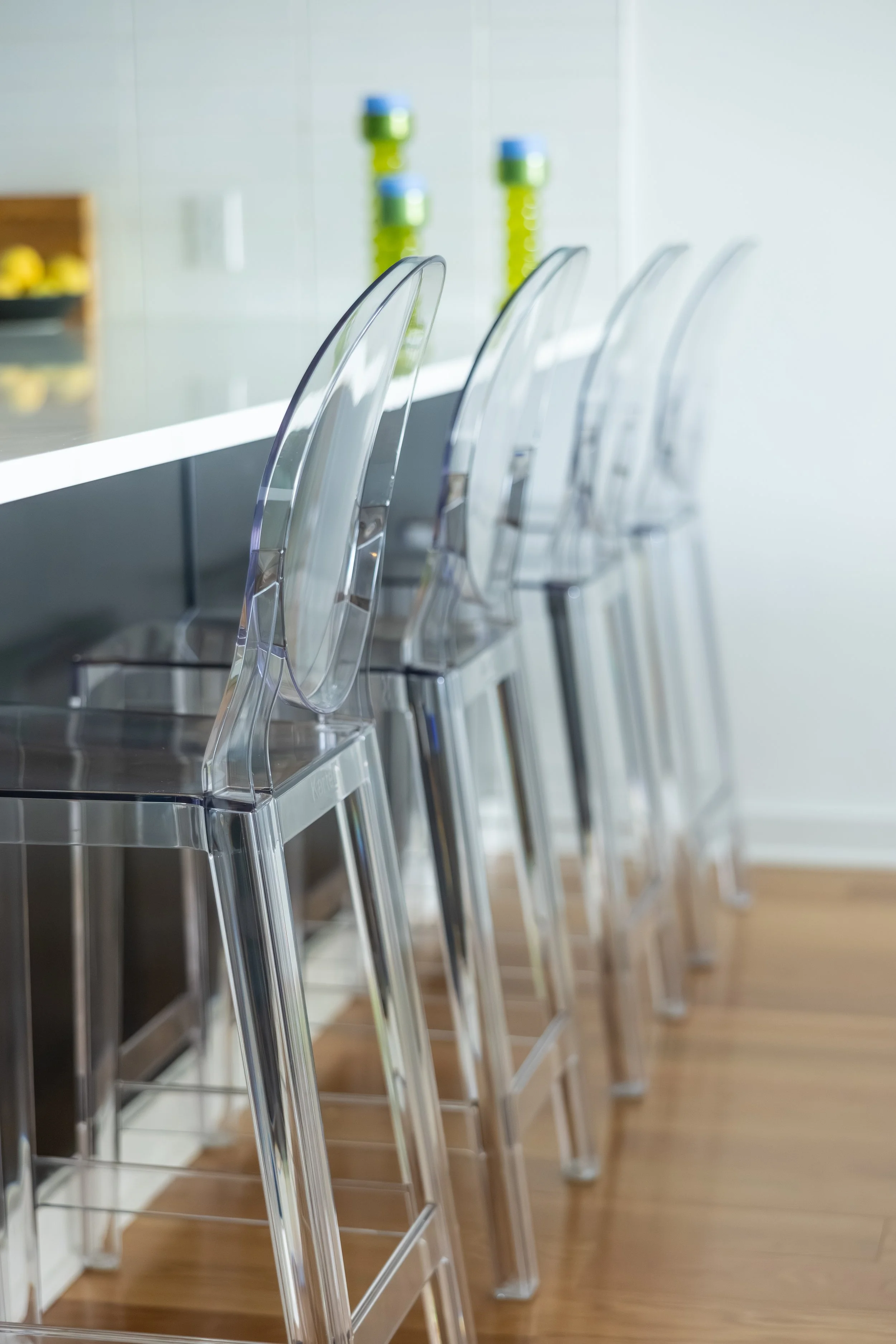 A row of clear, modern barstools with transparent backs and metal legs lined up along a counter in a bright kitchen or dining area.