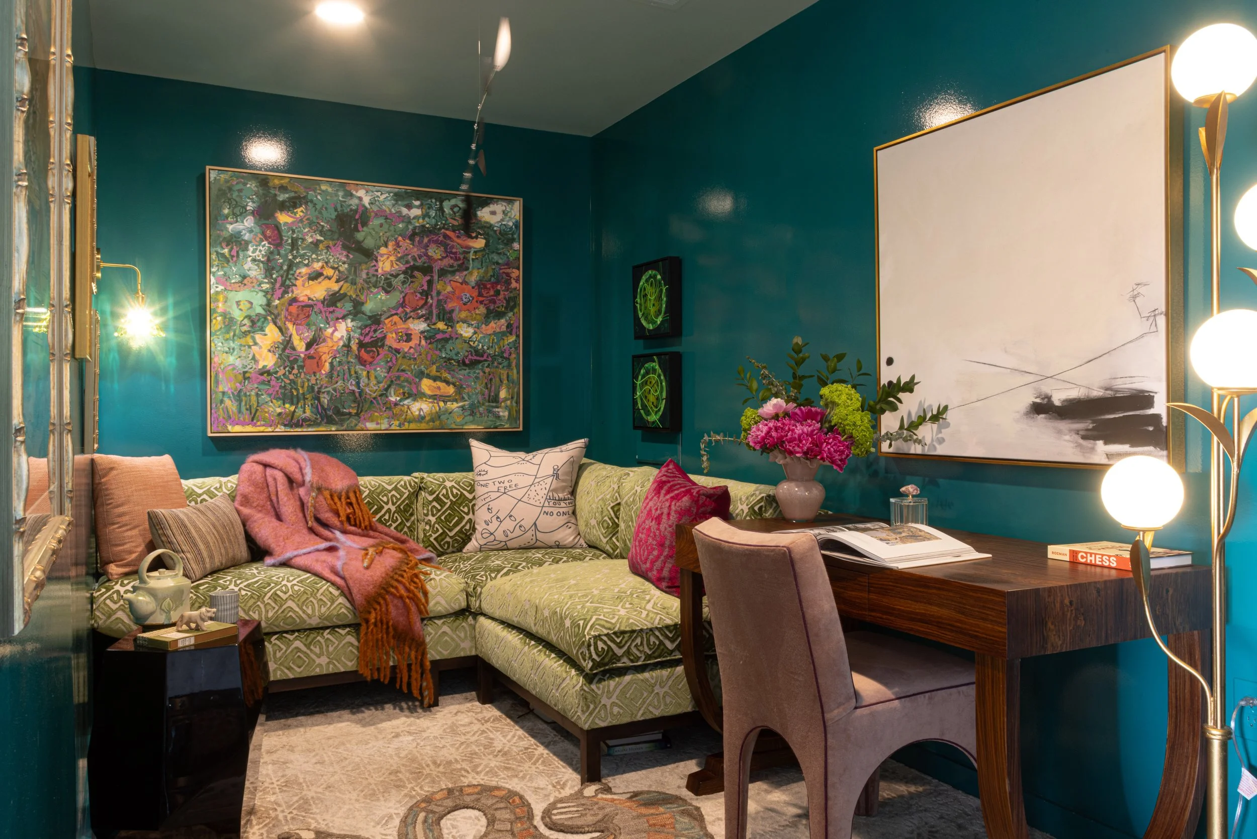 A cozy living room with teal walls, decorated with colorful abstract art and two framed green artwork. There is a green patterned sectional sofa adorned with multiple pillows and a pink throw blanket. A flower arrangement with pink and green flowers sits on a wooden table, along with a book on chess. To the right, a modern floor lamp with round white bulbs provides lighting. Part of a gold-framed mirror and a teal cabinet are visible on the sides.