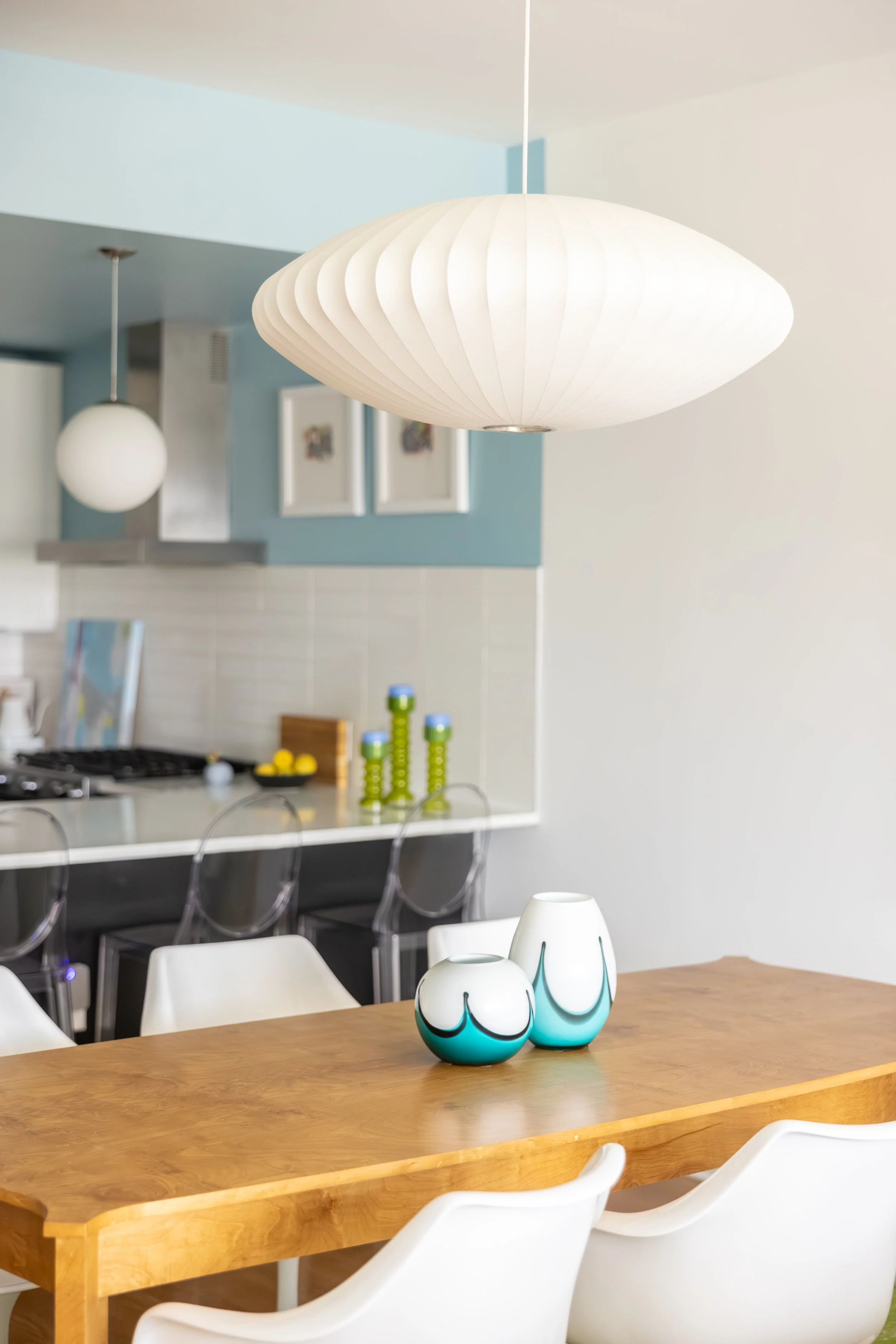 Modern kitchen and dining area with wooden table and white chairs, decorative vases, hanging pendant lights, and a blue accent wall with framed artwork.