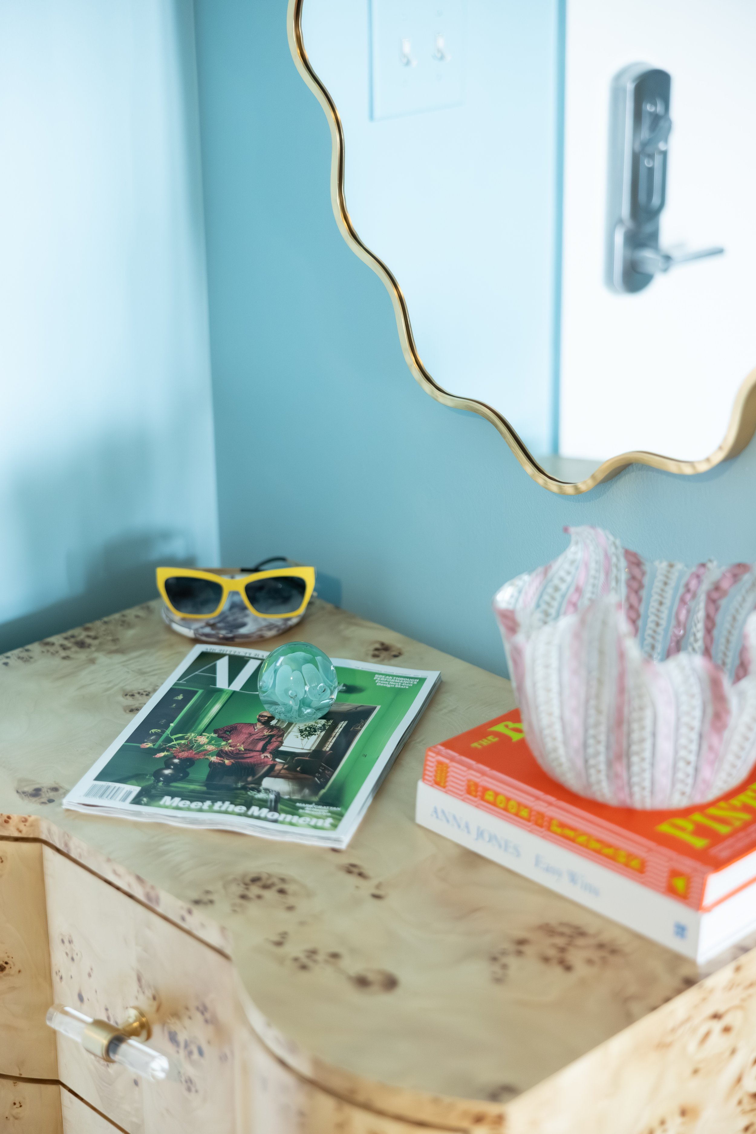 A marble tabletop with a magazine titled "Meet the Moment," a pair of yellow sunglasses, a glass paperweight, and a small wrapped gift. In the background, a mirror with a gold-wavy border hangs on a light blue wall, with a reflection of a door handle. Part of a striped fabric bag and a stack of books are also visible.