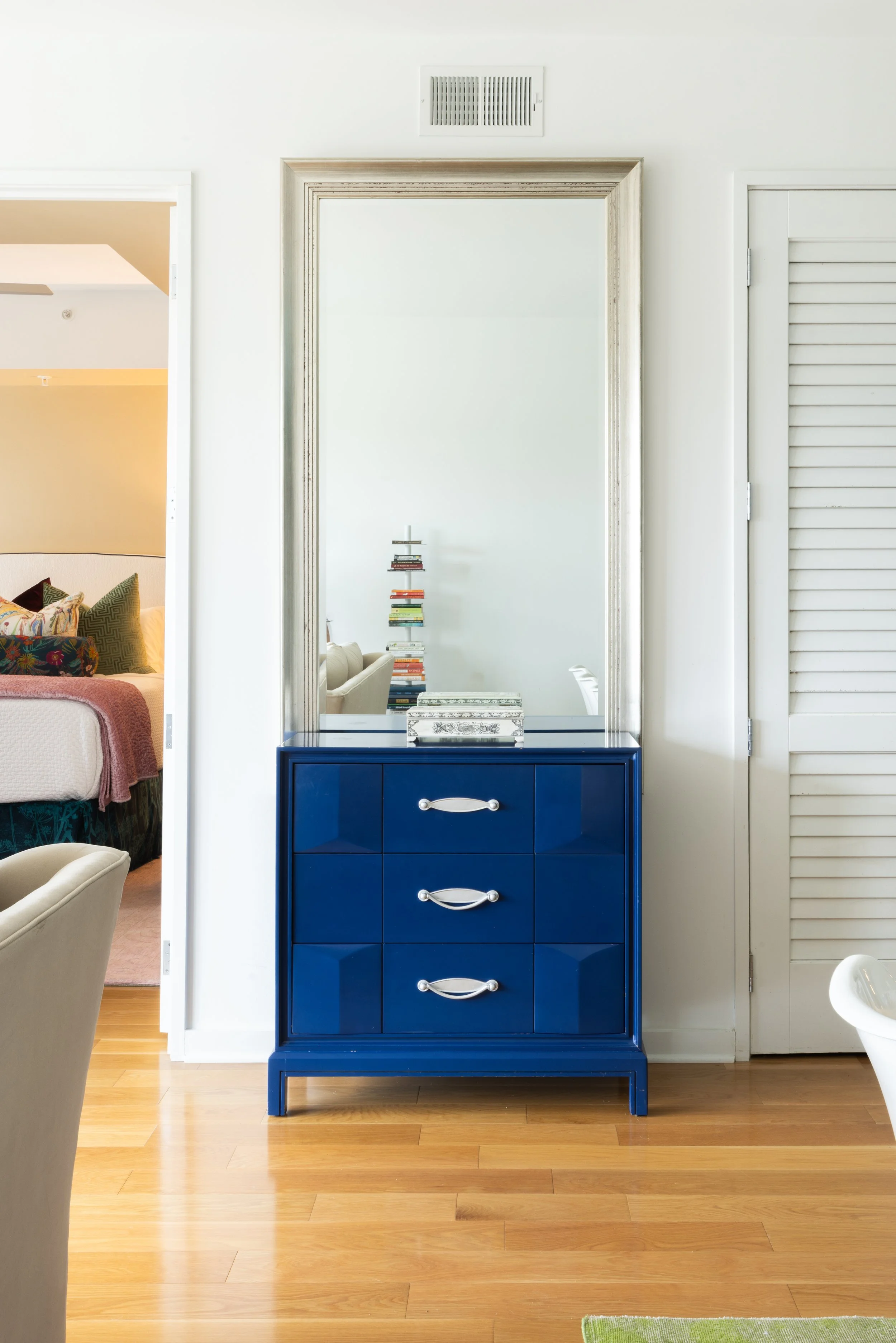 A blue dresser with six drawers and silver handles standing against a white wall in a room, with a large mirror behind it reflecting a stack of books and part of a living room.
