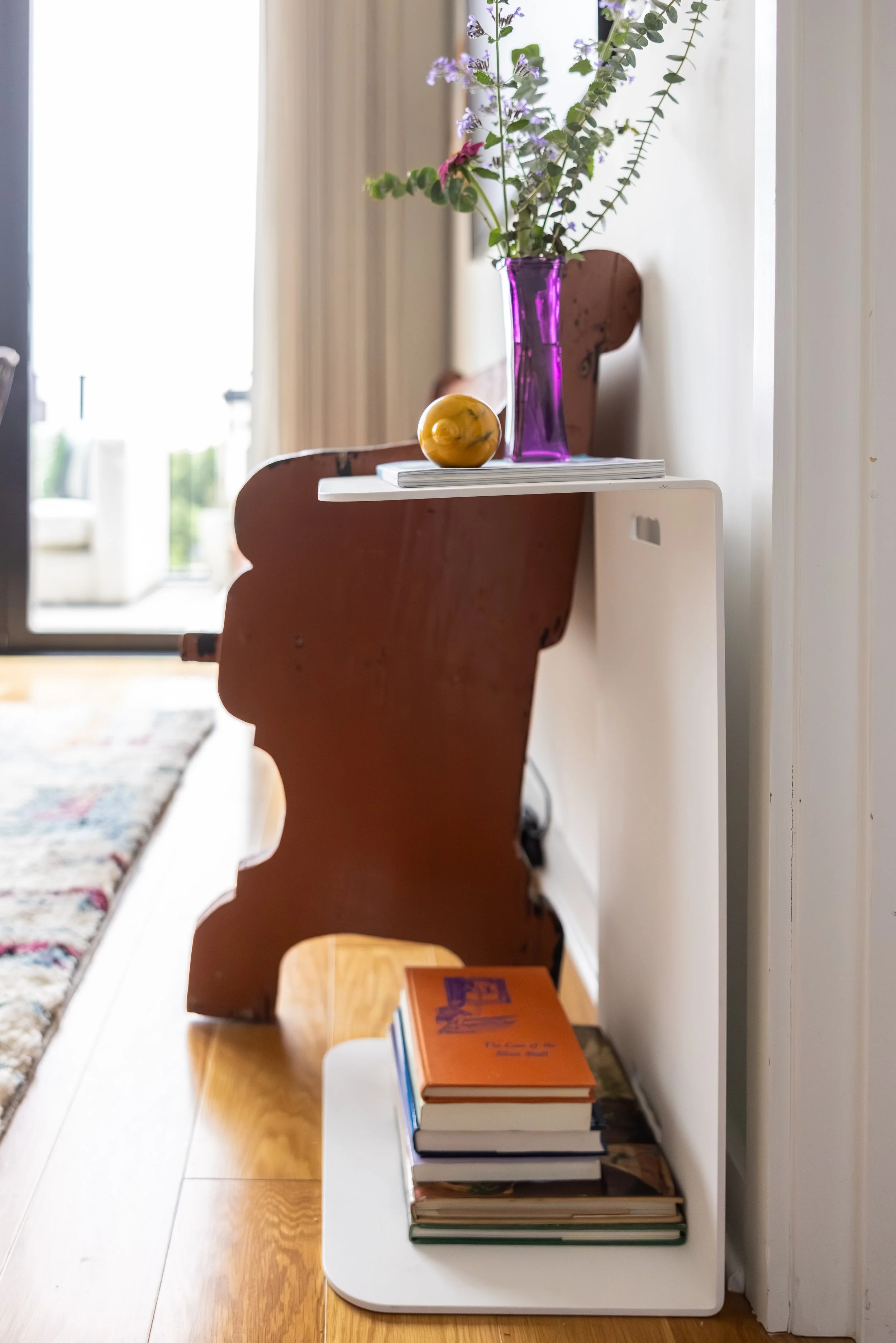 A white shelf against a wall holding a stack of books with an orange book on top, a purple vase with flowers, and a small yellow decorative object.