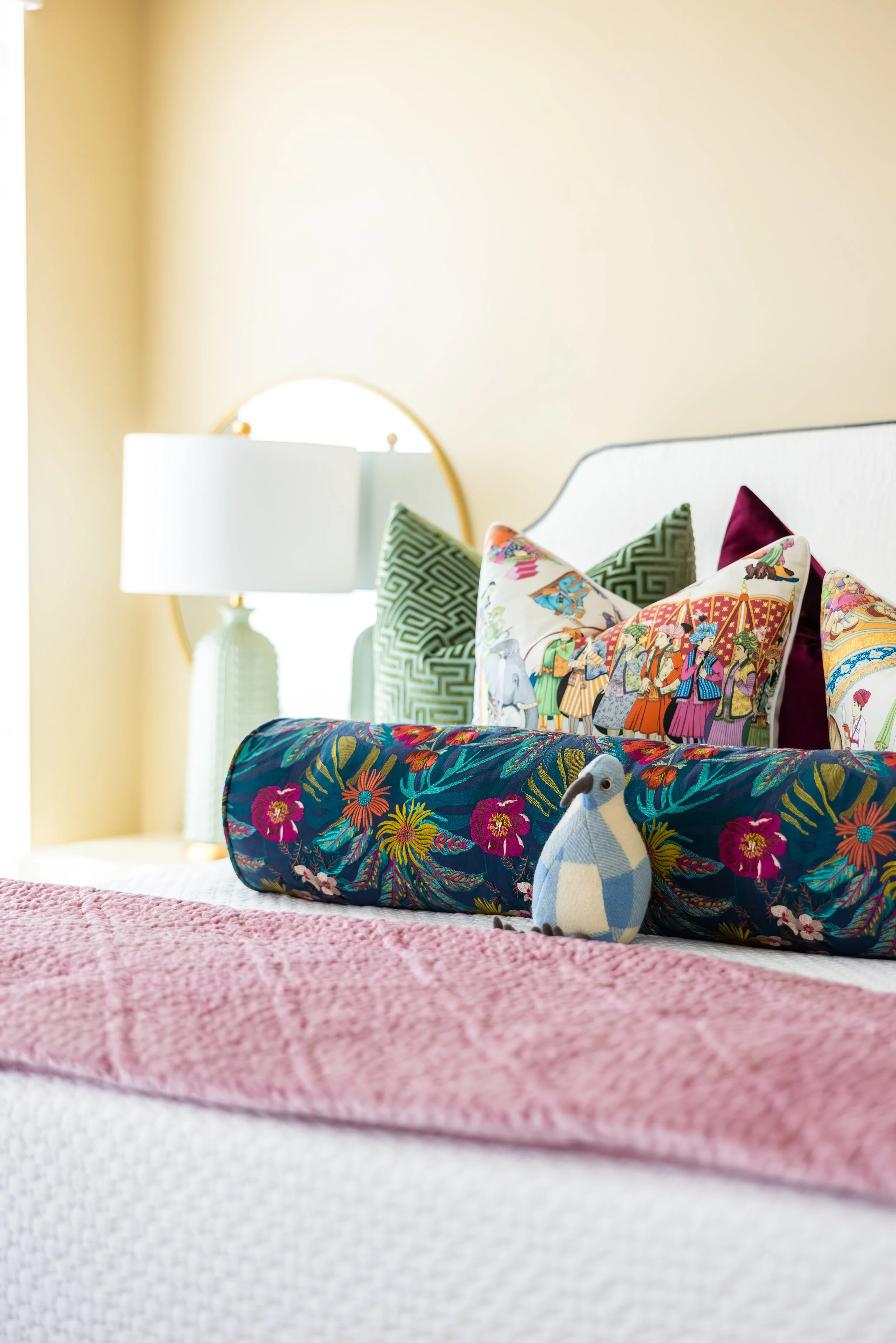 A neatly made bed with a pink textured blanket at the foot. The headboard has colorful pillows with Asian-inspired designs, including green geometric patterns and a multicolored tapestry pillow. On the left side, there is a white lamp with a textured base on a nightstand, and a bright window with a curved gold-framed mirror behind it in the background.