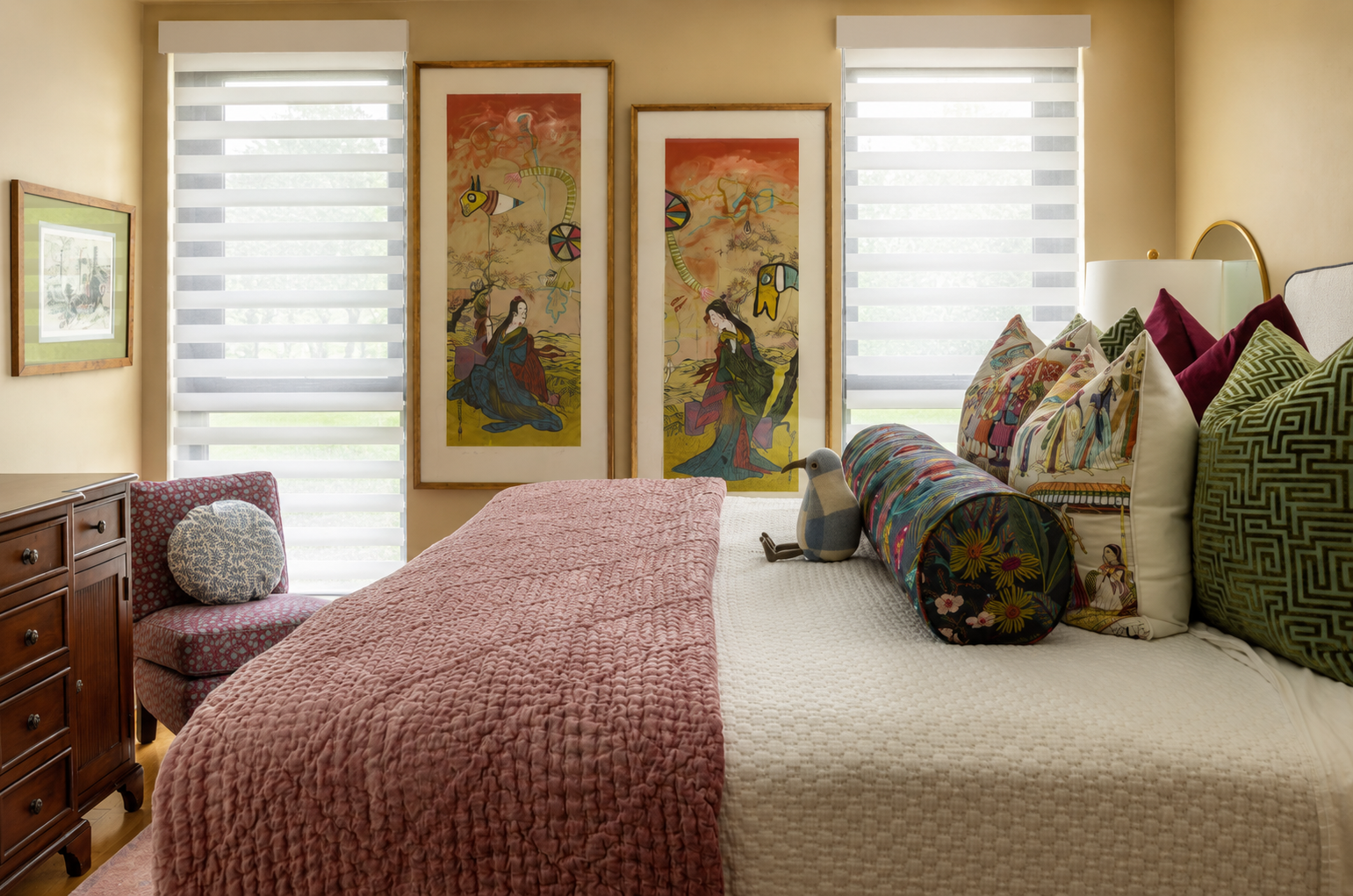 A bedroom with a bed covered in a pink quilt, decorated with colorful pillows and a fabric dog plush, and flanked by two large windows with striped shades, with framed artwork on the walls.