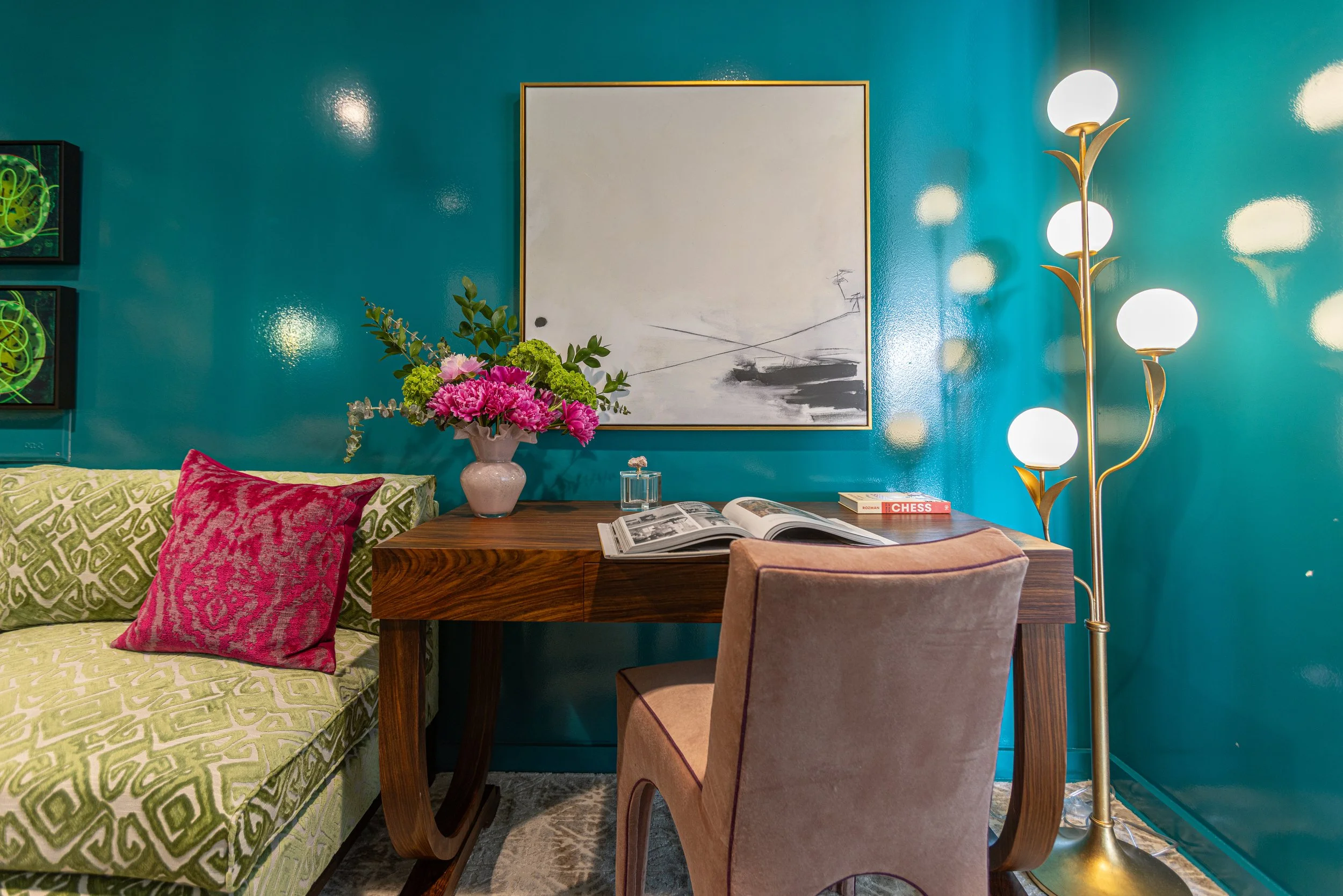 A living room with a teal wall, a wooden table, a pink flower arrangement, an open book, a pink pillow on a patterned green and yellow sofa, a modern lamp with white globes, and a large abstract black and white painting.