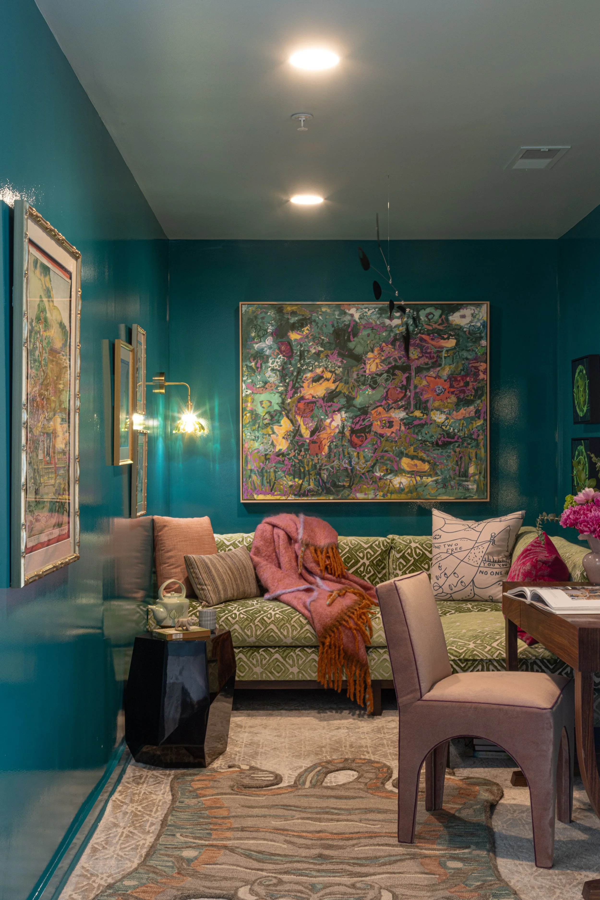 Living room with teal walls, framed artwork, and decorative pillows. A sofa with a pink blanket, contemporary side table, and a purple armchair. Ceiling lights, wall-mounted lamp, and a patterned area rug are visible.