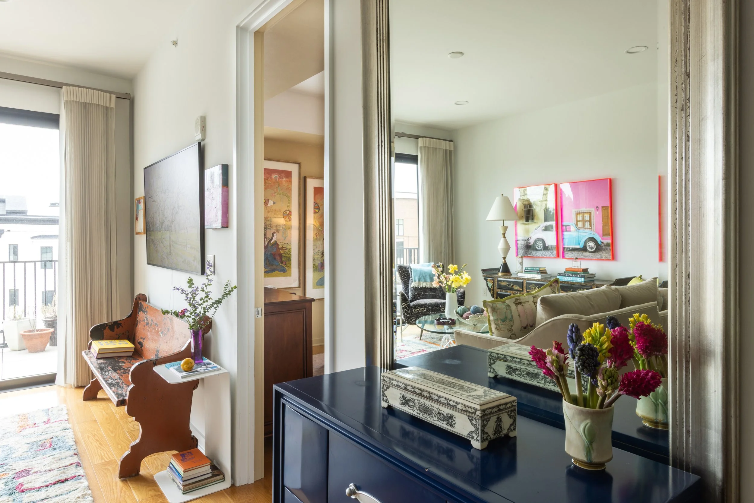 Living room with artwork on the walls, a black cabinet with a floral box and vase of flowers, a beige couch, and mirrors reflecting a pink building and cars outside